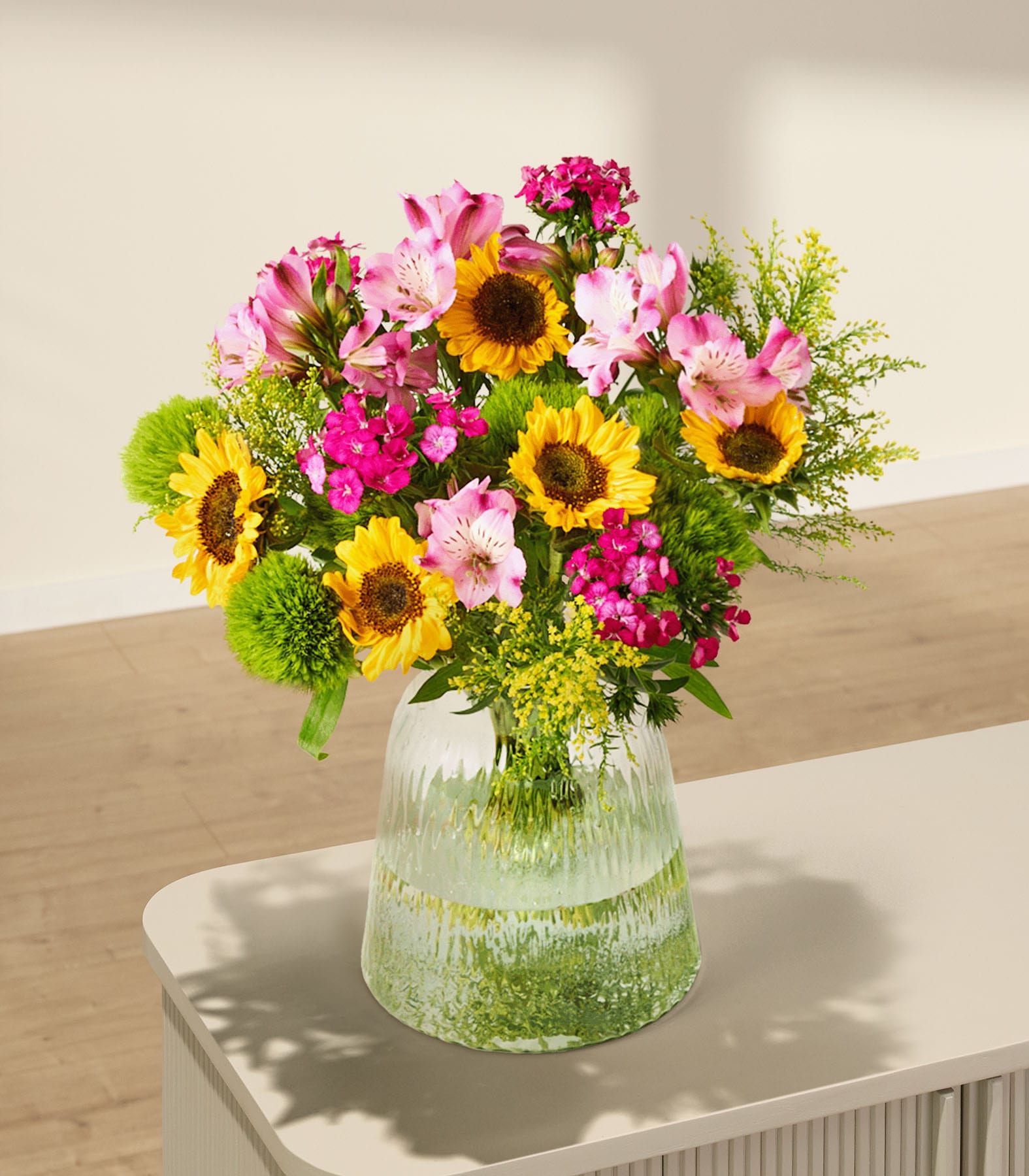 Ein leuchtender Strauß bunter Blumen, darunter Sonnenblumen sowie rosa und rote Blüten, wird vor einem schlichten weißen Hintergrund in den Händen einer Person gehalten.