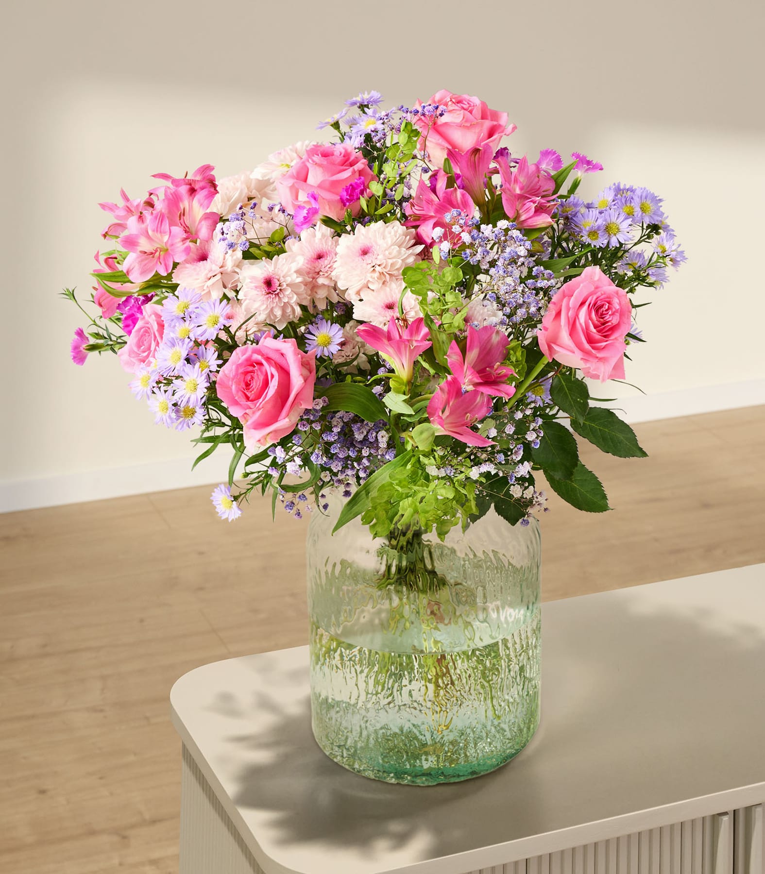 Ein farbenfroher Strauß aus rosa Rosen, lila Blumen und Grünpflanzen wird in einer Glasvase vor einem schlichten weißen Hintergrund präsentiert.