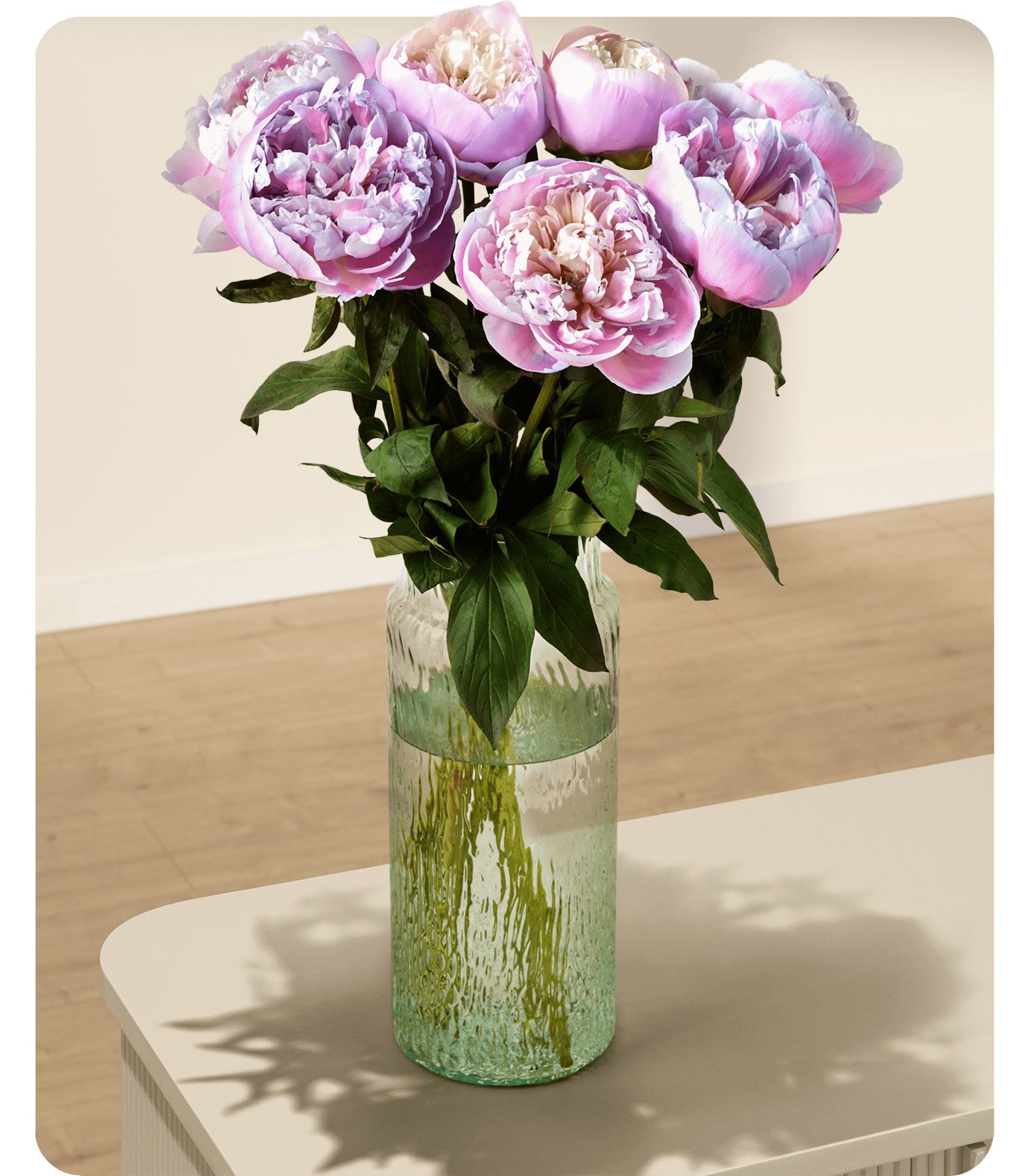 Ein Strauß üppiger, rosa Pfingstrosen in einer grünen Glasvase steht auf einer hellen Fläche vor einem schlichten Hintergrund.