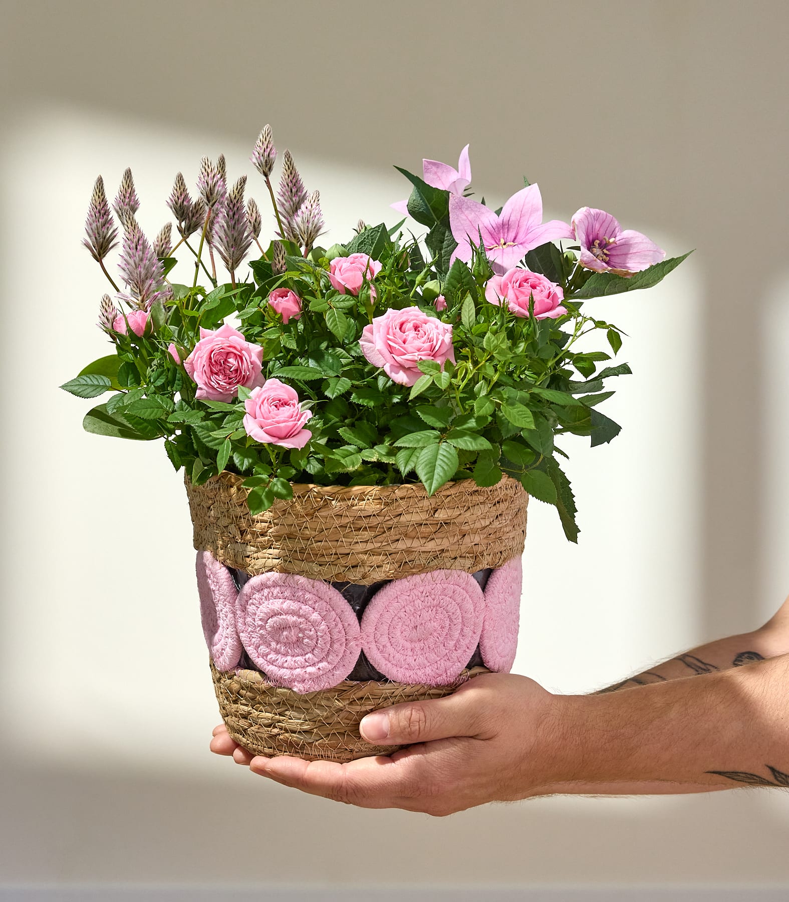 Ein lebendiges Arrangement aus rosa Rosen, Lilien und anderen Blumen in einem geflochtenen Korb vor schlichtem weißen Hintergrund.