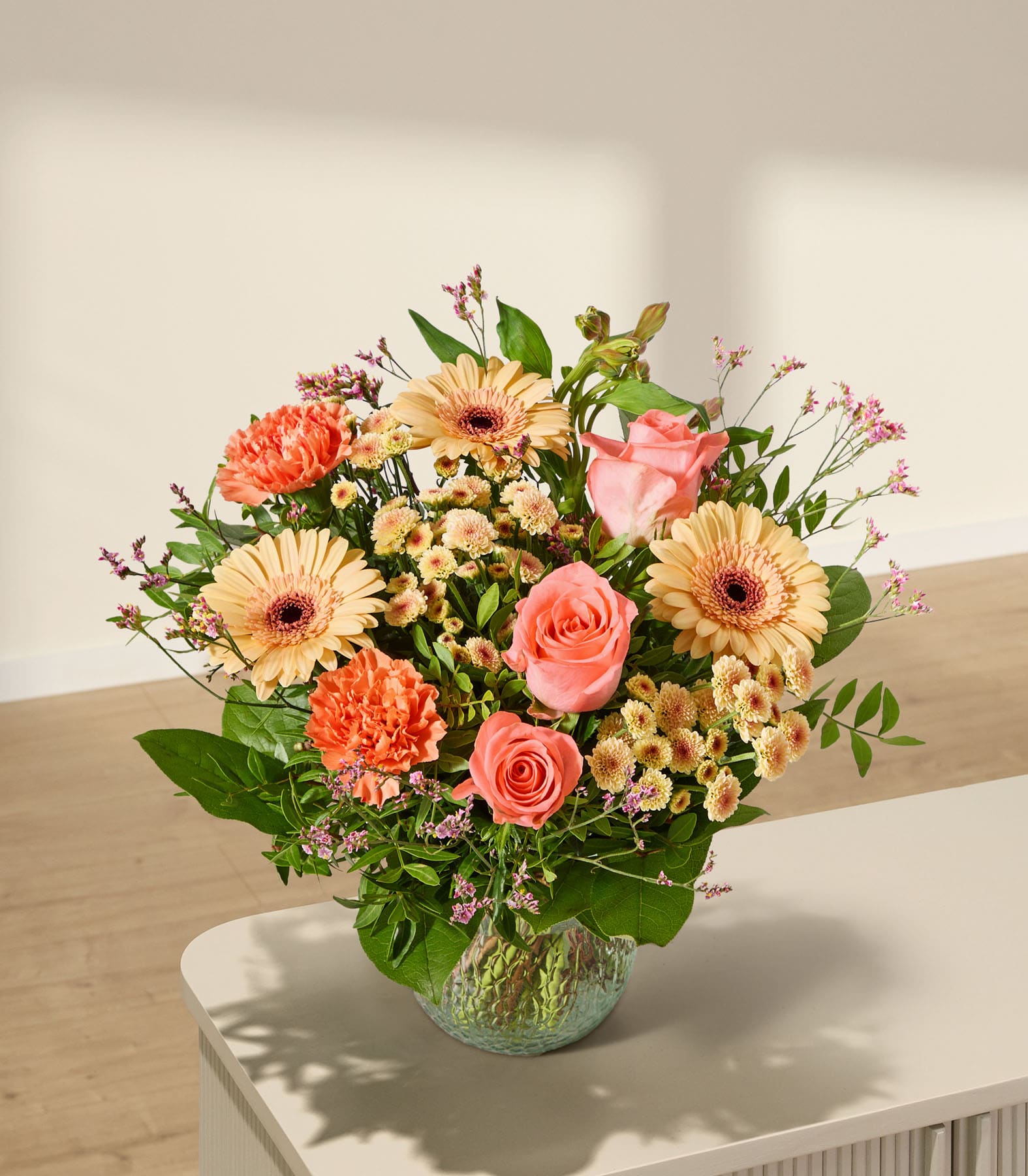 Ein leuchtender Strauß rosa und orangefarbener Blumen steht in einer strukturierten grünen Vase auf einer hellen Oberfläche, während im Hintergrund sanfte Schatten auf eine helle Wand fallen.
