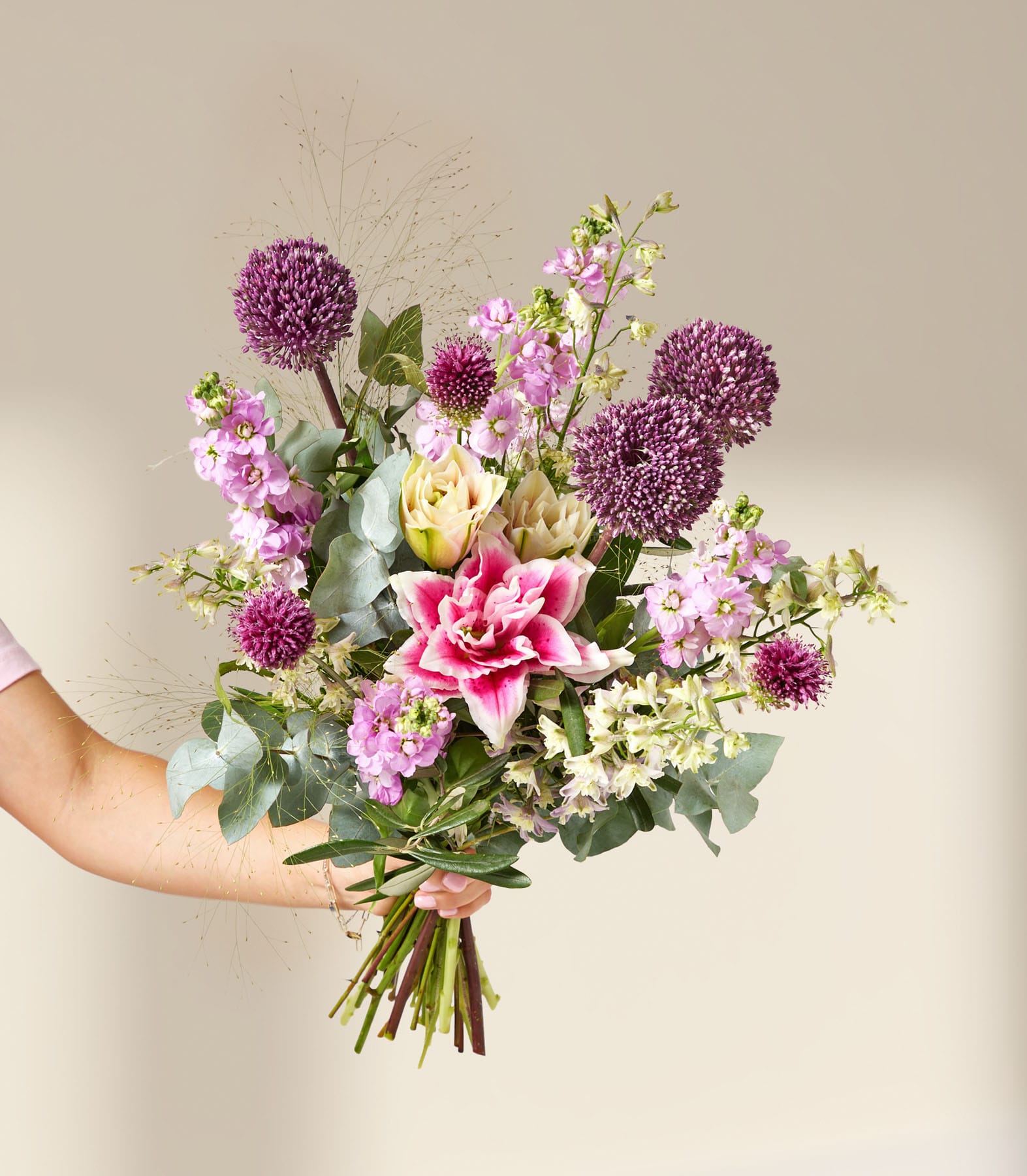 Eine Hand hält einen leuchtenden Strauß aus violettem Zierlauch, rosa Lilien und zarten weißen Blüten vor einem sanften, neutralen Hintergrund.
