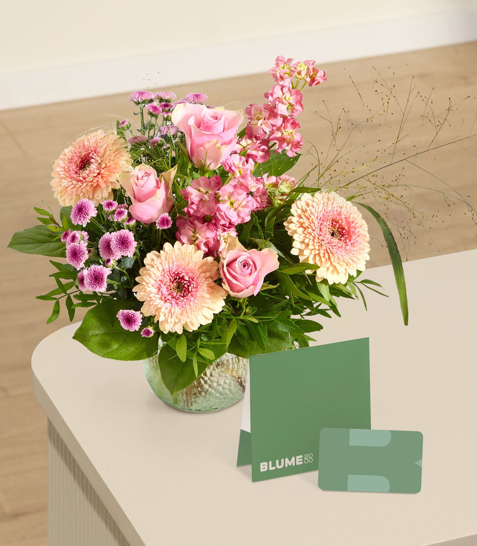 Ein leuchtender Strauß rosa und pfirsichfarbener Blumen steht in einer Glasvase auf einem hellen Tisch, davor liegen eine grüne Karte und eine Geschenkkarte, vor einem verschwommenen Hintergrund eines Zimmers.