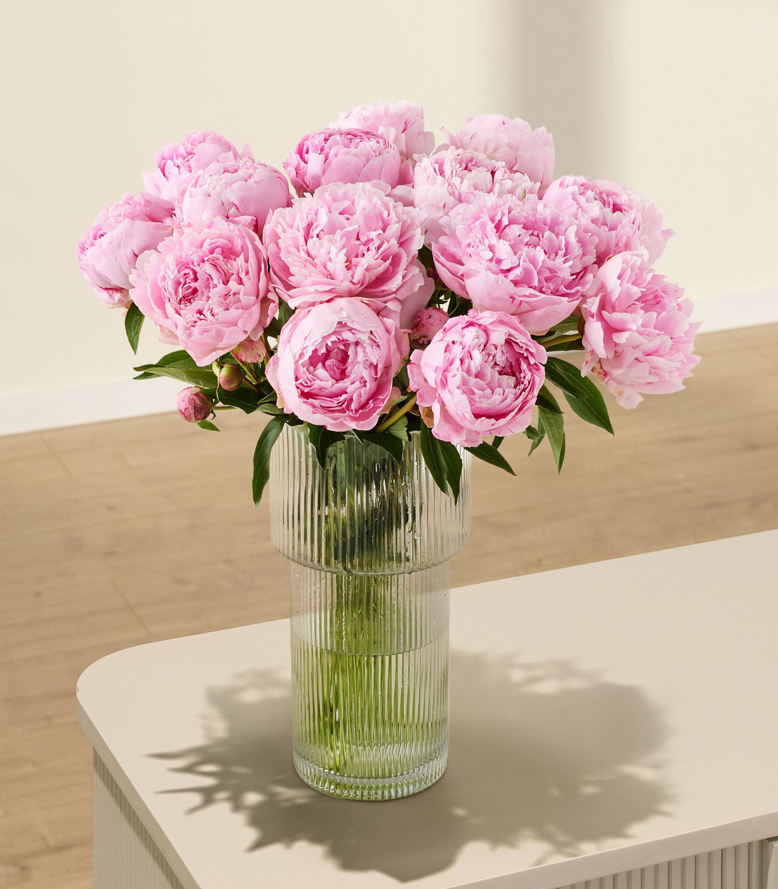 Ein Strauß üppiger, rosa Pfingstrosen in einer Glasvase steht auf einer Holzoberfläche vor einem schlichten, beigen Hintergrund.