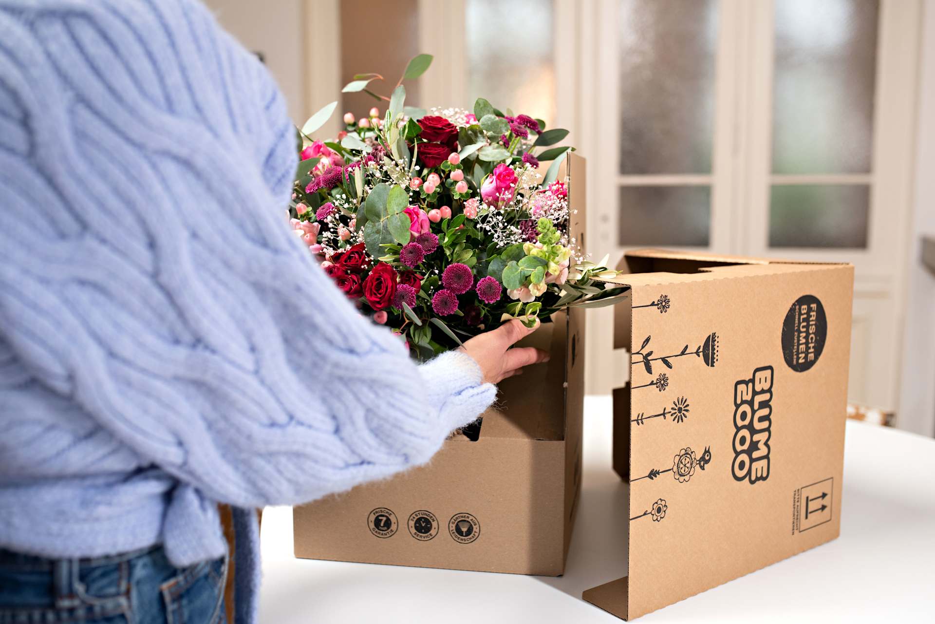Eine Person in einem blauen Pullover hält einen Strauß bunter Blumen vor einem Karton mit Versandetiketten.