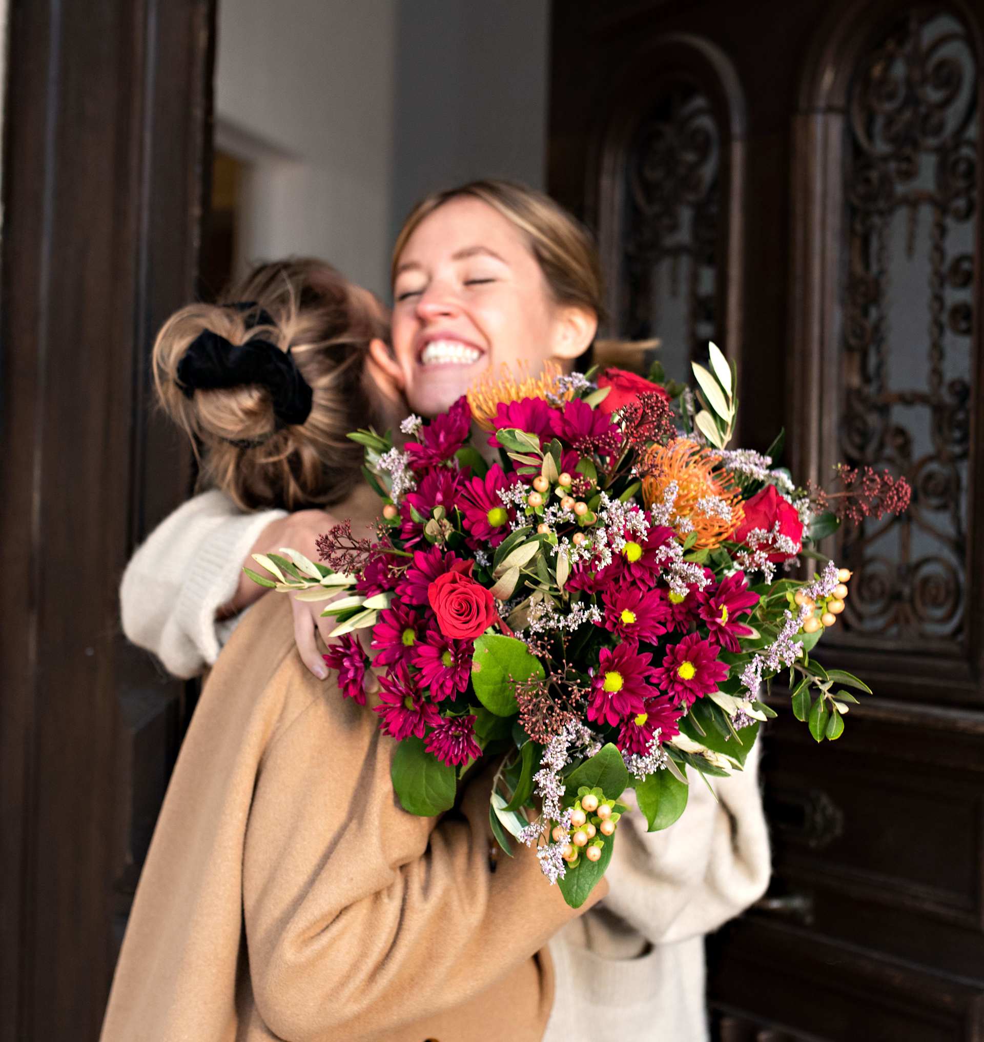 Eine lächelnde Frau in einem beigen Mantel hält einen farbenfrohen Strauß bunter Blumen vor dem Hintergrund einer verzierten Holztür.