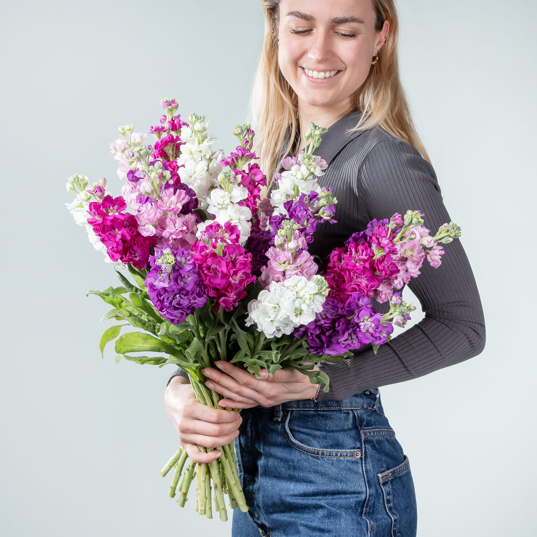 Eine lächelnde junge Frau hält einen großen Strauß leuchtend rosa, violetter und weißer Blumen vor einem schlichten hellen Hintergrund.