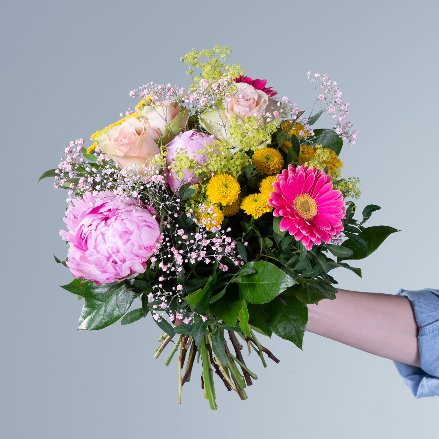 Ein lebendiger Strauß bunter Blumen, darunter rosa Pfingstrosen, gelbe Gerbera und weißes Schleierkraut, wird vor einem schlichten grauen Hintergrund in den Händen einer Person gehalten.
