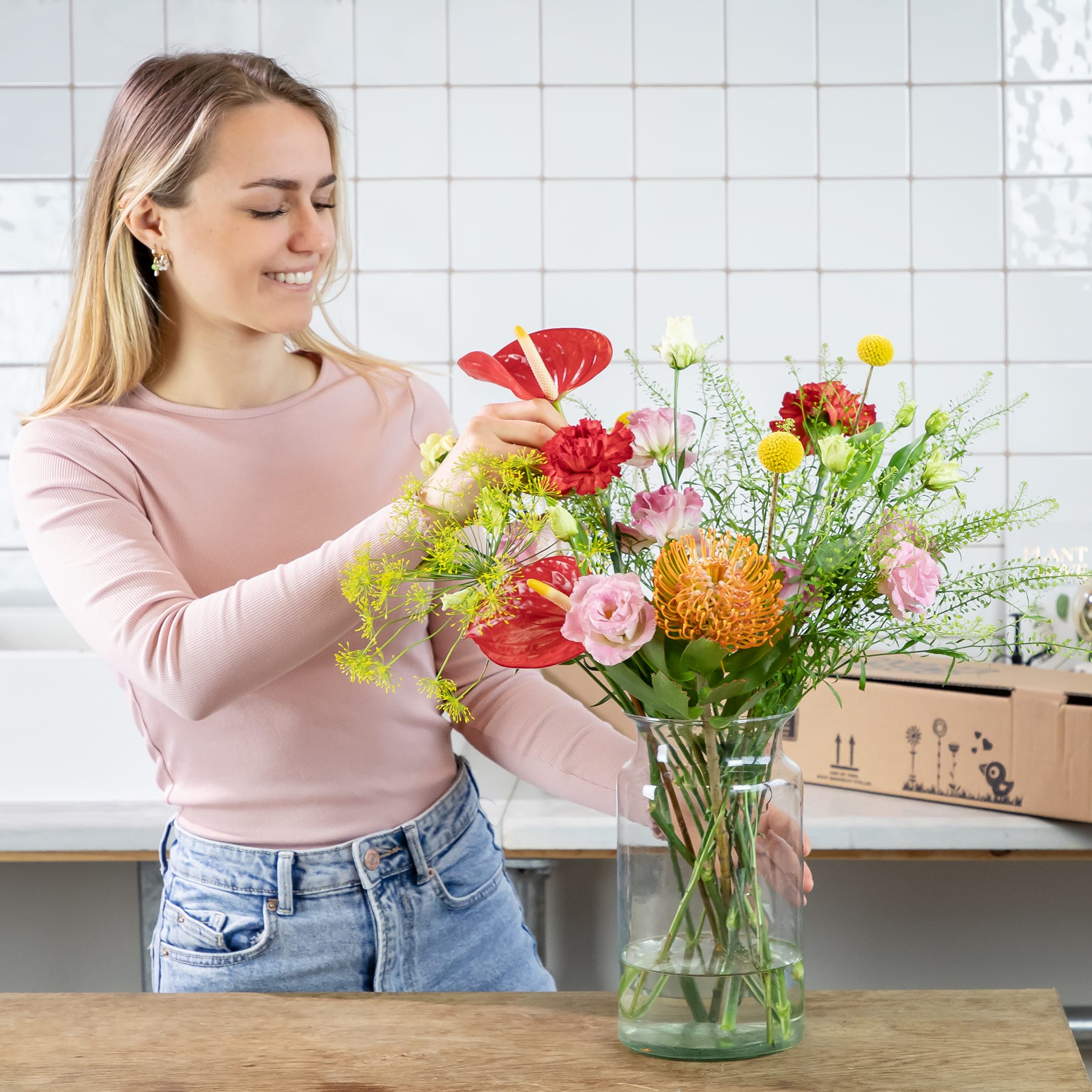 Eine junge Frau mit blondem Haar arrangiert einen farbenfrohen Strauß bunter Blumen in einer Glasvase auf einer Küchentheke, im Hintergrund ist eine geflieste Wand zu sehen.