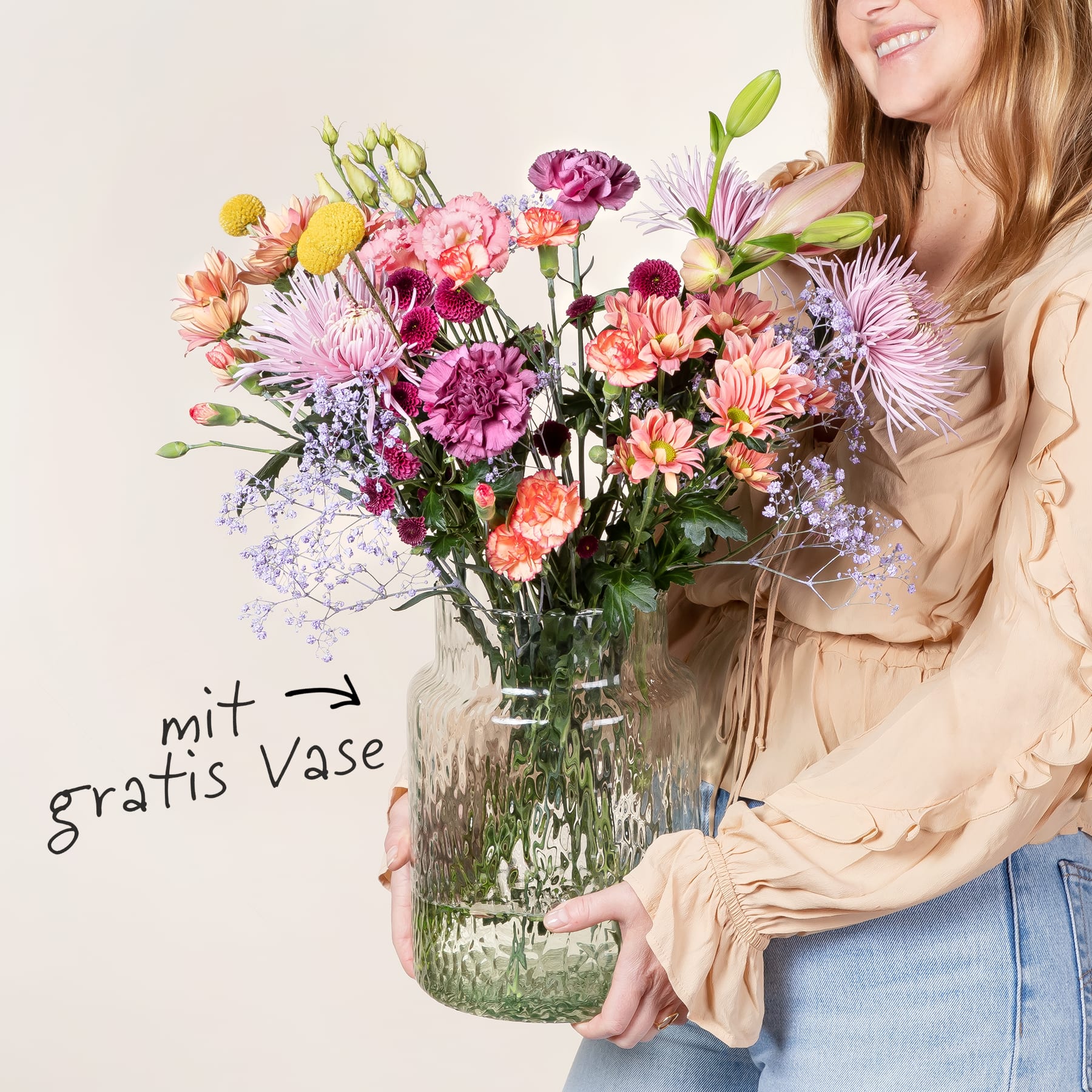 Ein leuchtender Strauß bunter Blumen, darunter rosa Nelken, violette Dahlien und gelbe Narzissen, wird von einer lächelnden Frau in einem beigen Pullover vor einem schlichten Hintergrund gehalten.