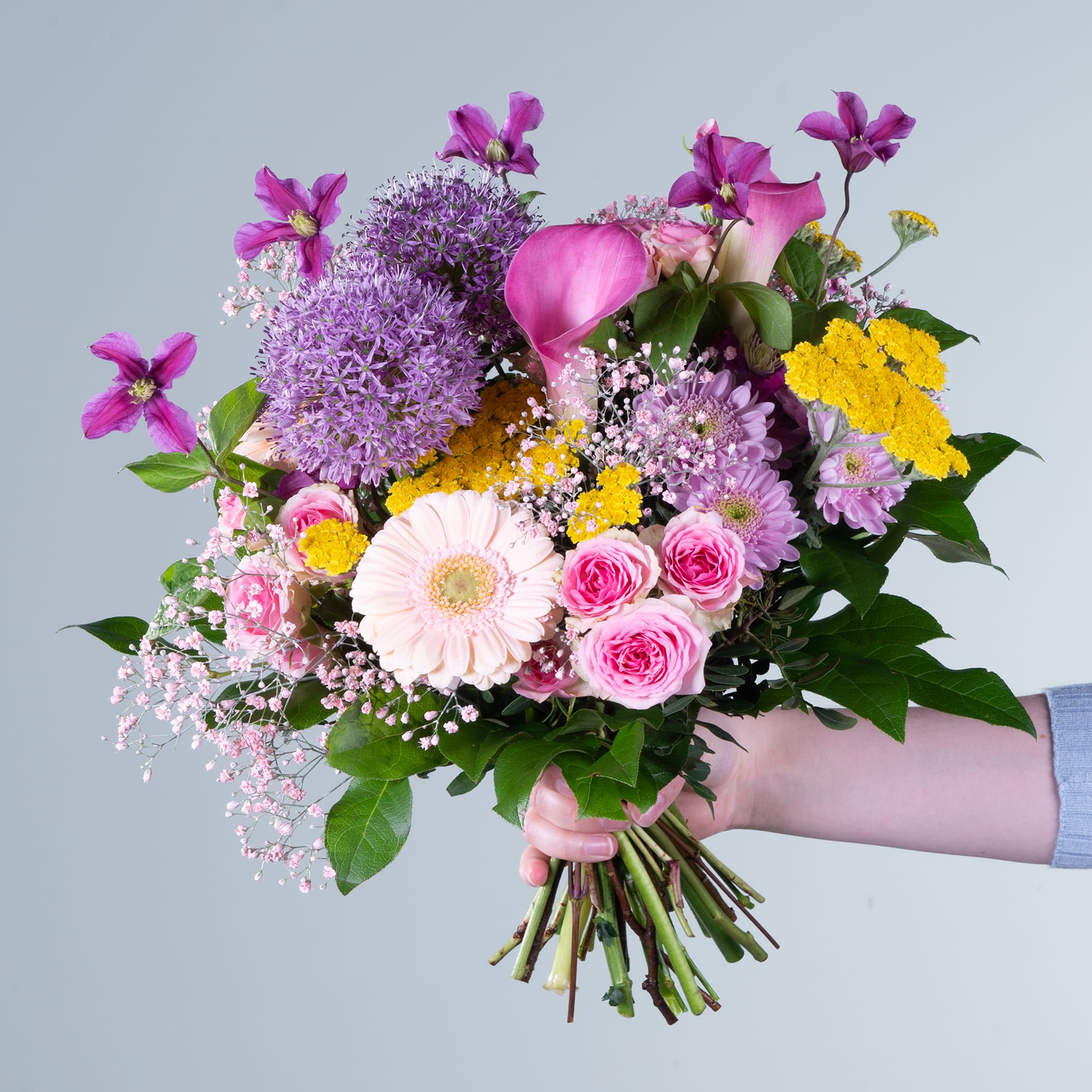 Ein leuchtender Strauß verschiedener bunter Blumen, darunter rosa Rosen, gelbe Gänseblümchen und lila Allium, wird vor einem schlichten grauen Hintergrund in den Händen einer Person gehalten.