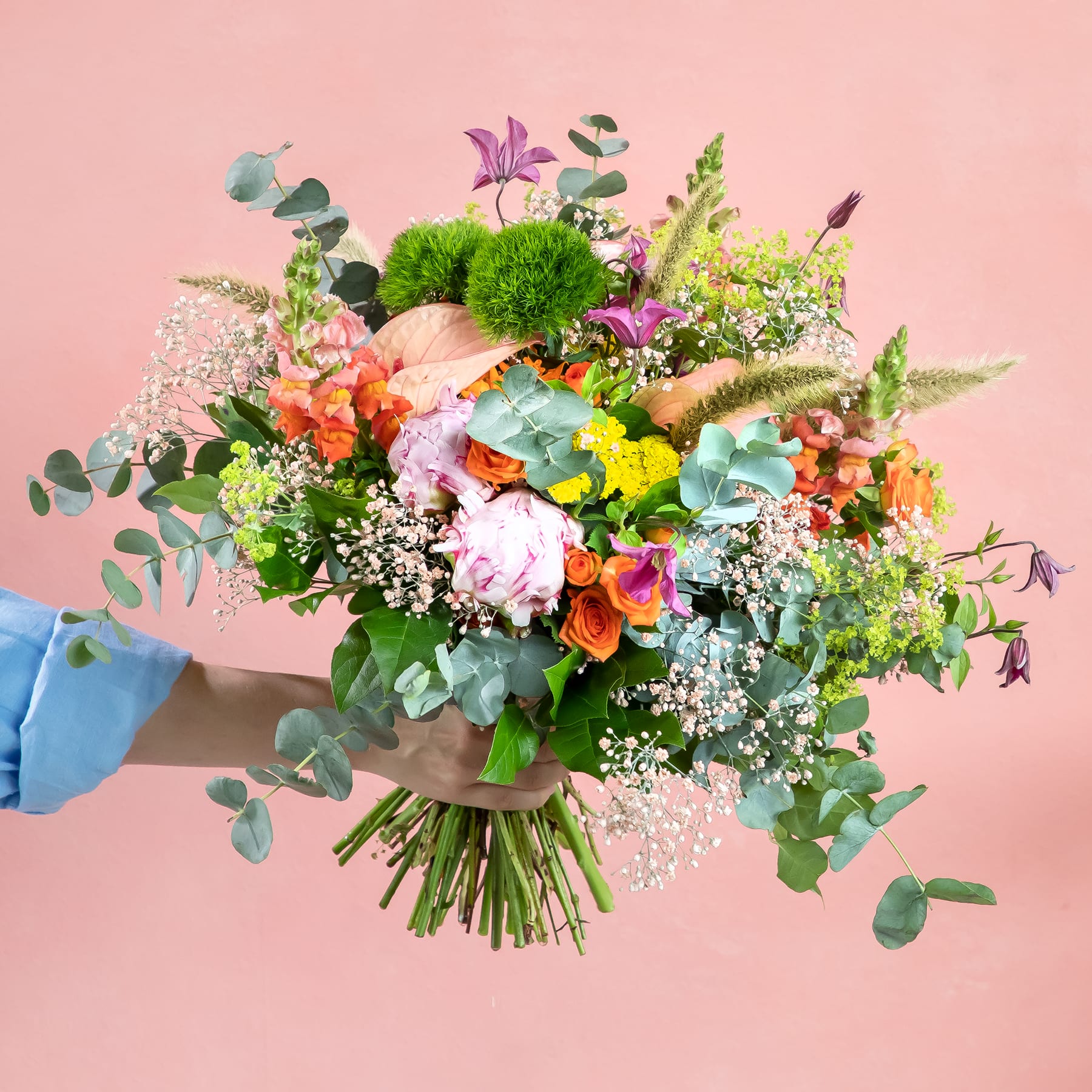 Ein lebendiger und farbenfroher Strauß verschiedener Blumen, darunter Rosen, Nelken und Eukalyptus, den eine Person vor einem zartrosa Hintergrund in den Händen hält.