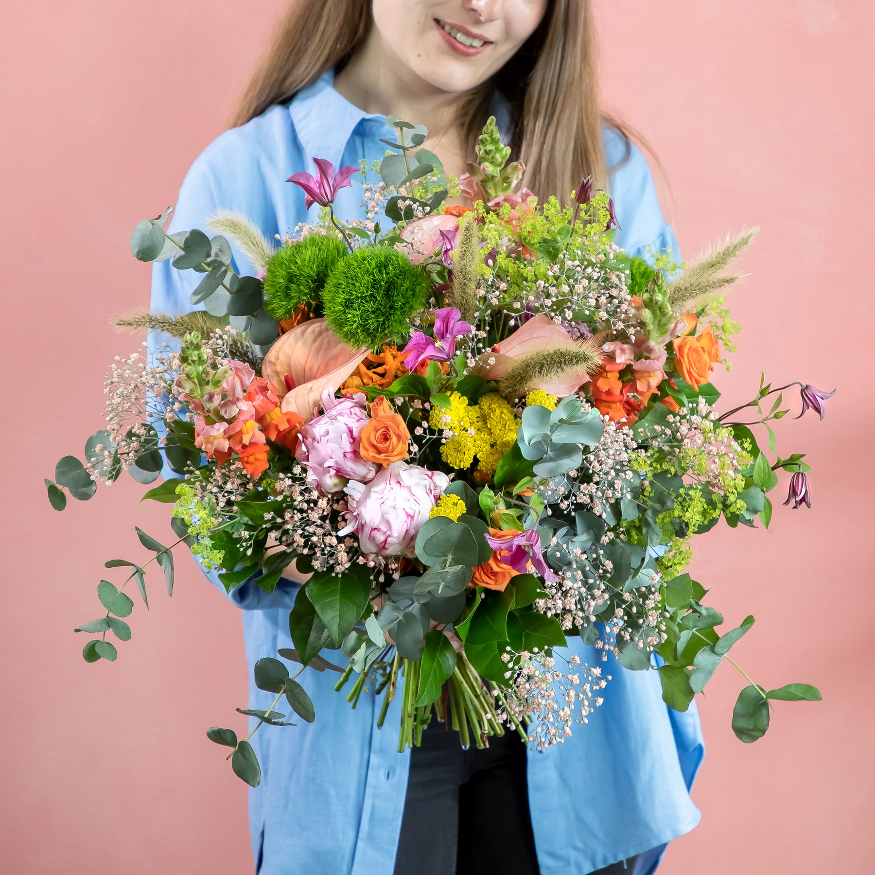Eine junge Frau in einem blauen Hemd hält vor einem schlichten rosa Hintergrund einen leuchtend bunten Blumenstrauß, darunter Rosen, Chrysanthemen und Grünpflanzen.