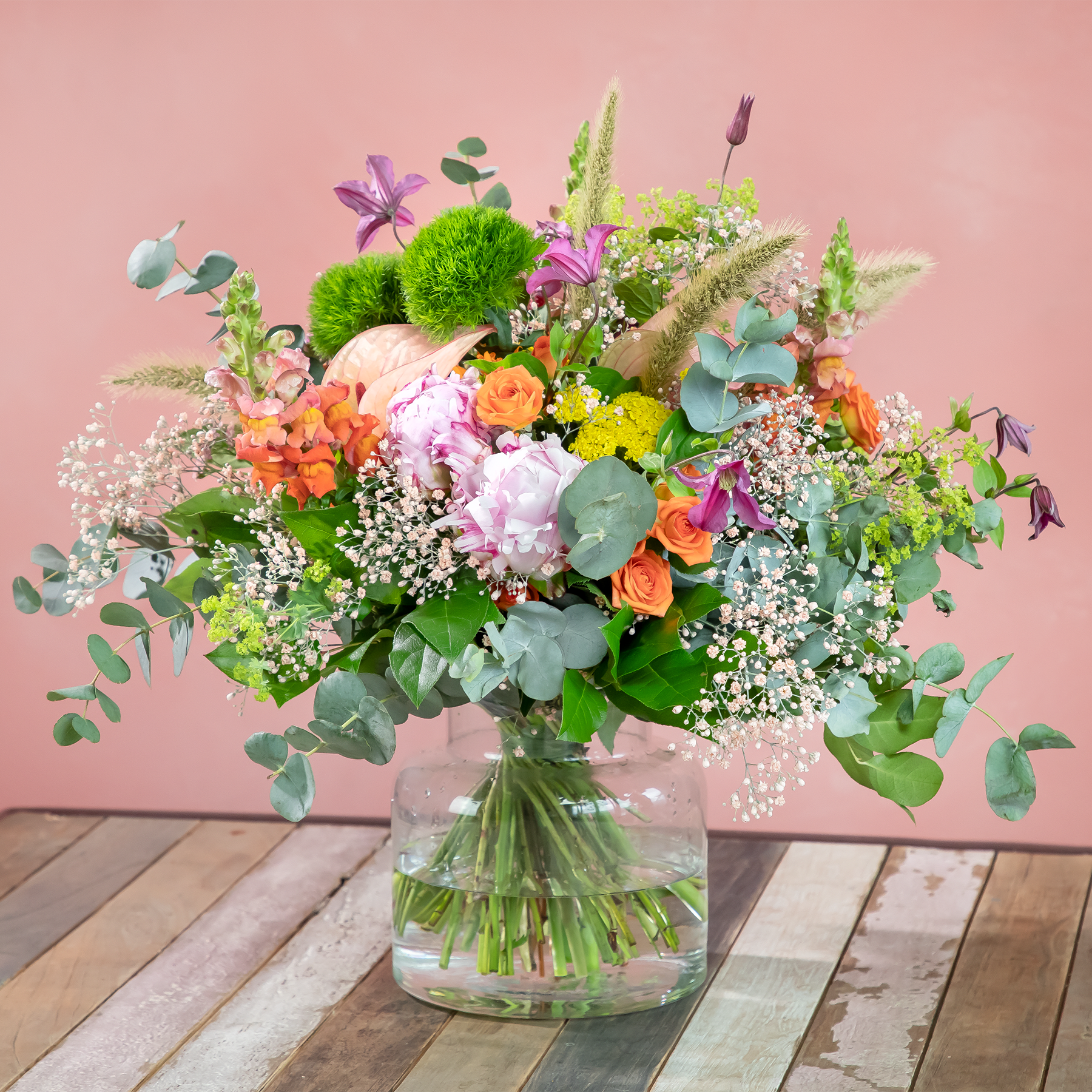 Ein lebendiger und üppiger Strauß verschiedener Blumen, darunter Pfingstrosen, Rosen und Eukalyptus, arrangiert in einer Glasvase vor einem zartrosa Hintergrund auf einer Holzoberfläche.