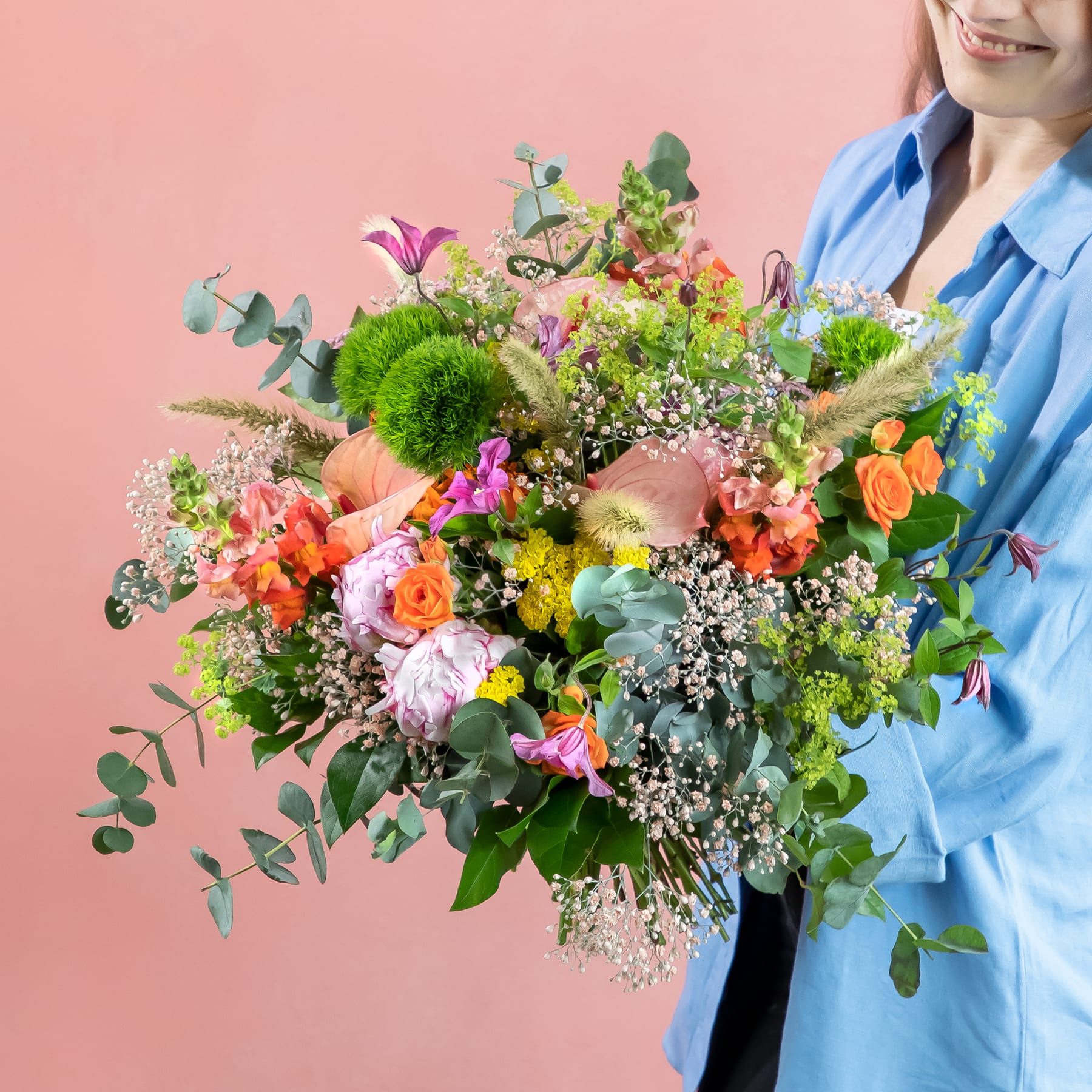 Ein lebendiger und üppiger Strauß bunter Blumen, darunter Rosen, Nelken und Grünpflanzen, gehalten von einer Person in einem blauen Hemd vor einem rosa Hintergrund.