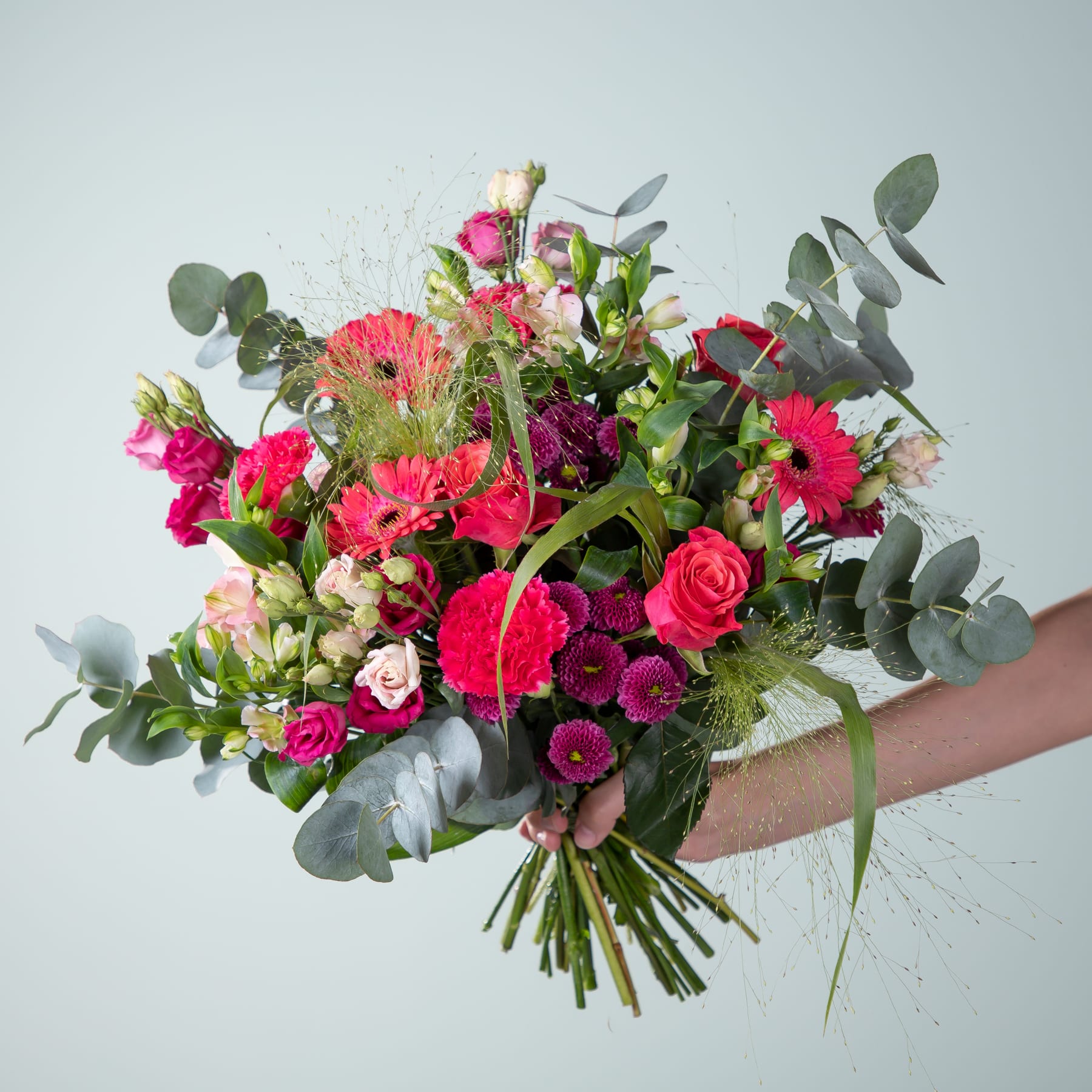 Ein lebendiger Strauß verschiedener bunter Blumen, darunter Rosen, Gerbera und Grünpflanzen, wird vor einem schlichten Hintergrund in den Händen einer Person gehalten.