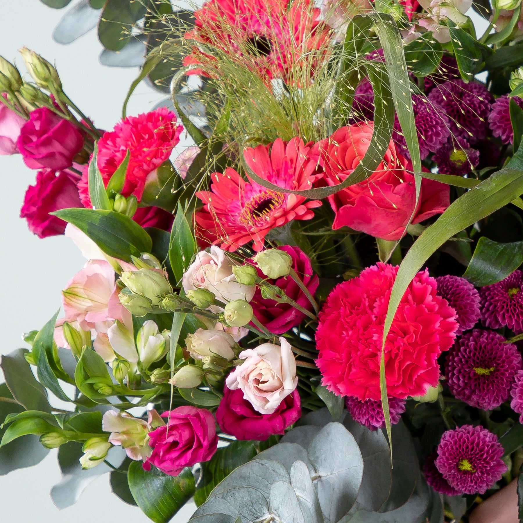 Ein lebendiger Strauß verschiedener bunter Blumen, darunter rote Nelken, rosa Rosen und lila Chrysanthemen, arrangiert vor einem hellen Hintergrund.