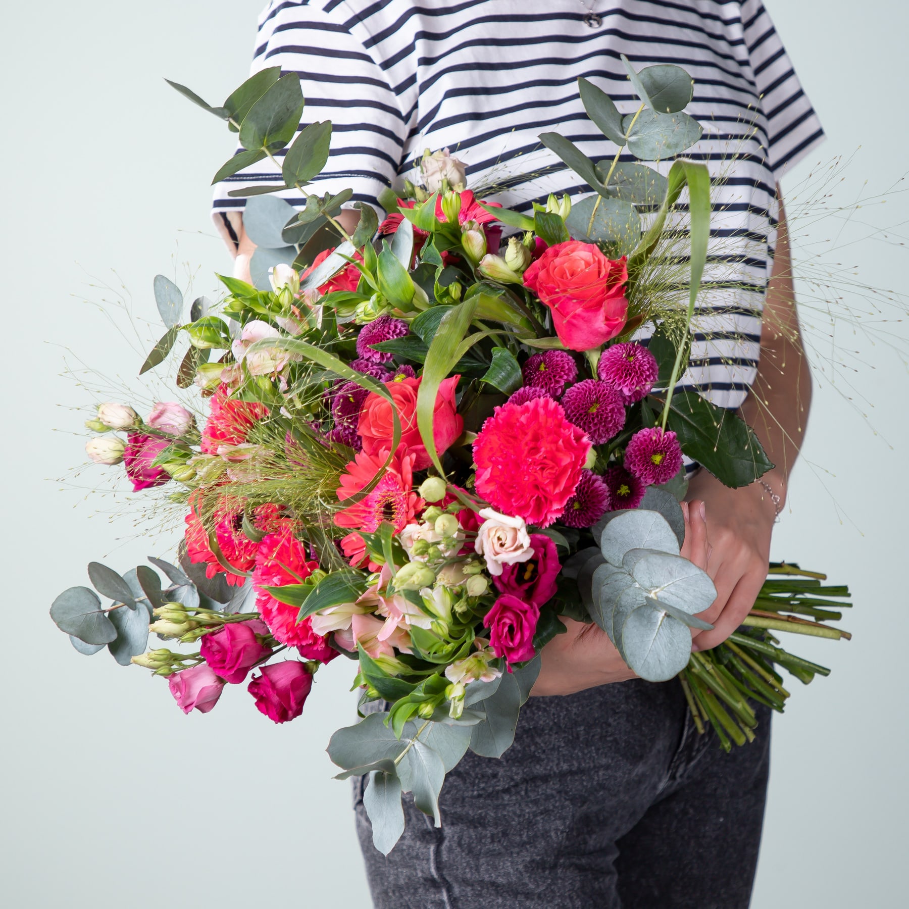 Ein leuchtender Strauß bunter Blumen, darunter rosa Nelken und Rosen, wird vor einer Person gehalten, die ein gestreiftes Hemd vor einem schlichten Hintergrund trägt.