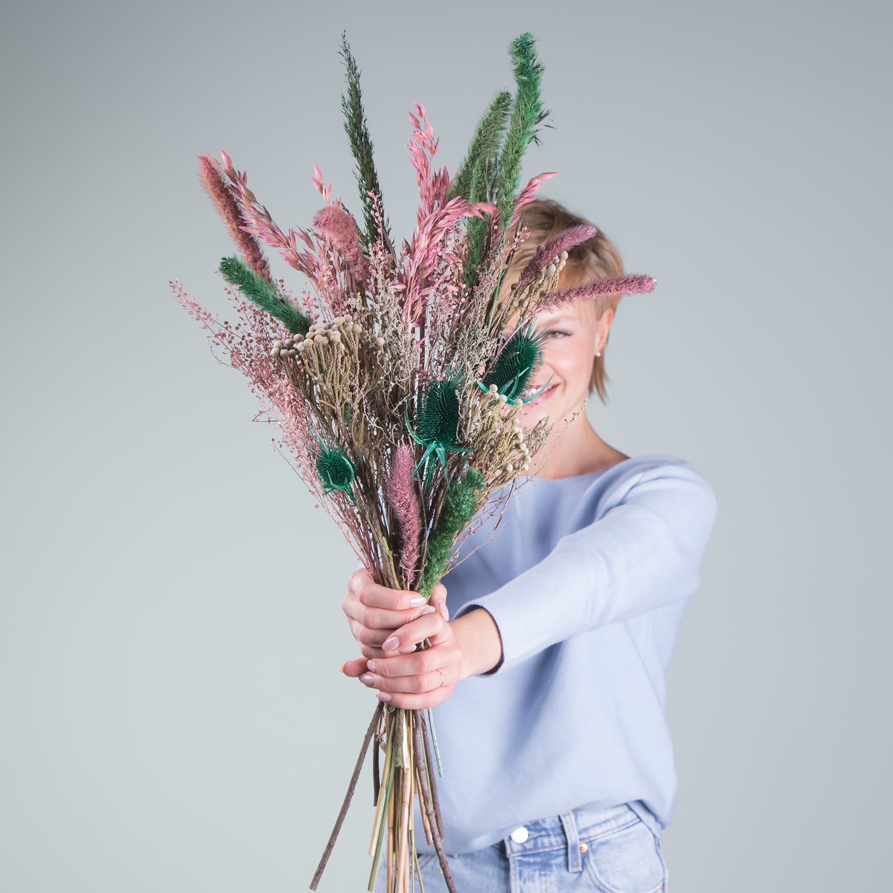 Eine Person hält einen großen Strauß lebendiger, bunter Trockenblumen und Pflanzen vor einem schlichten, hellen Hintergrund.
