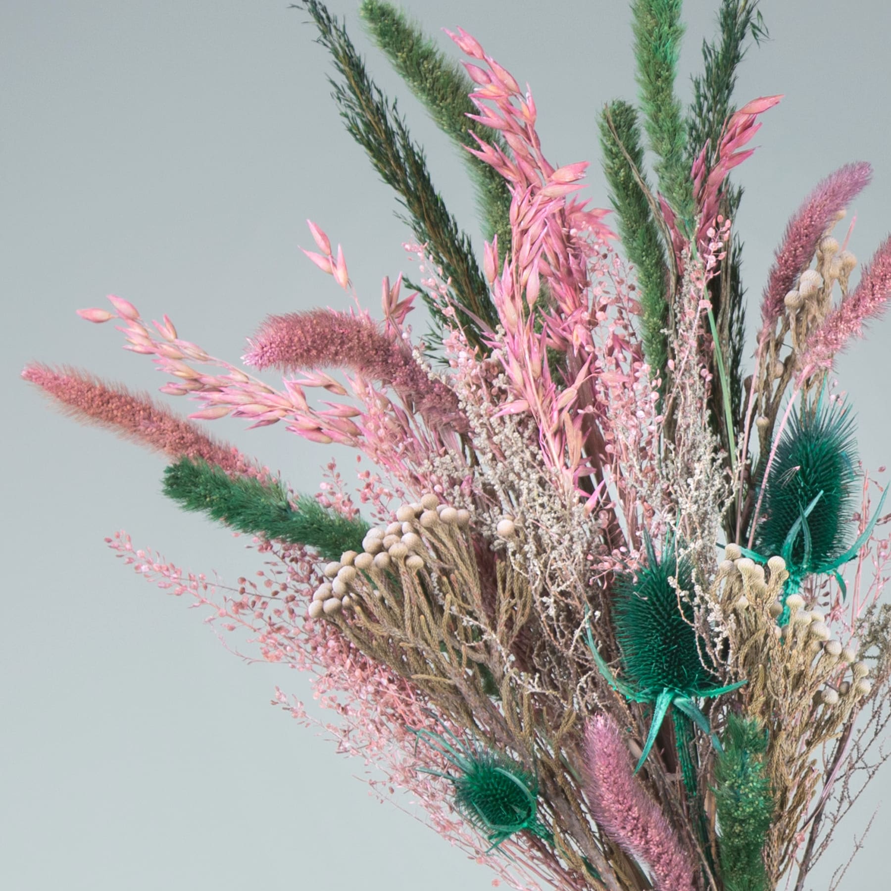 Ein lebendiger Strauß getrockneter und konservierter Blumen, darunter rosafarbene Federbüsche, grünes Laub und strukturierte Samenkapseln vor einem gedämpften grauen Hintergrund.