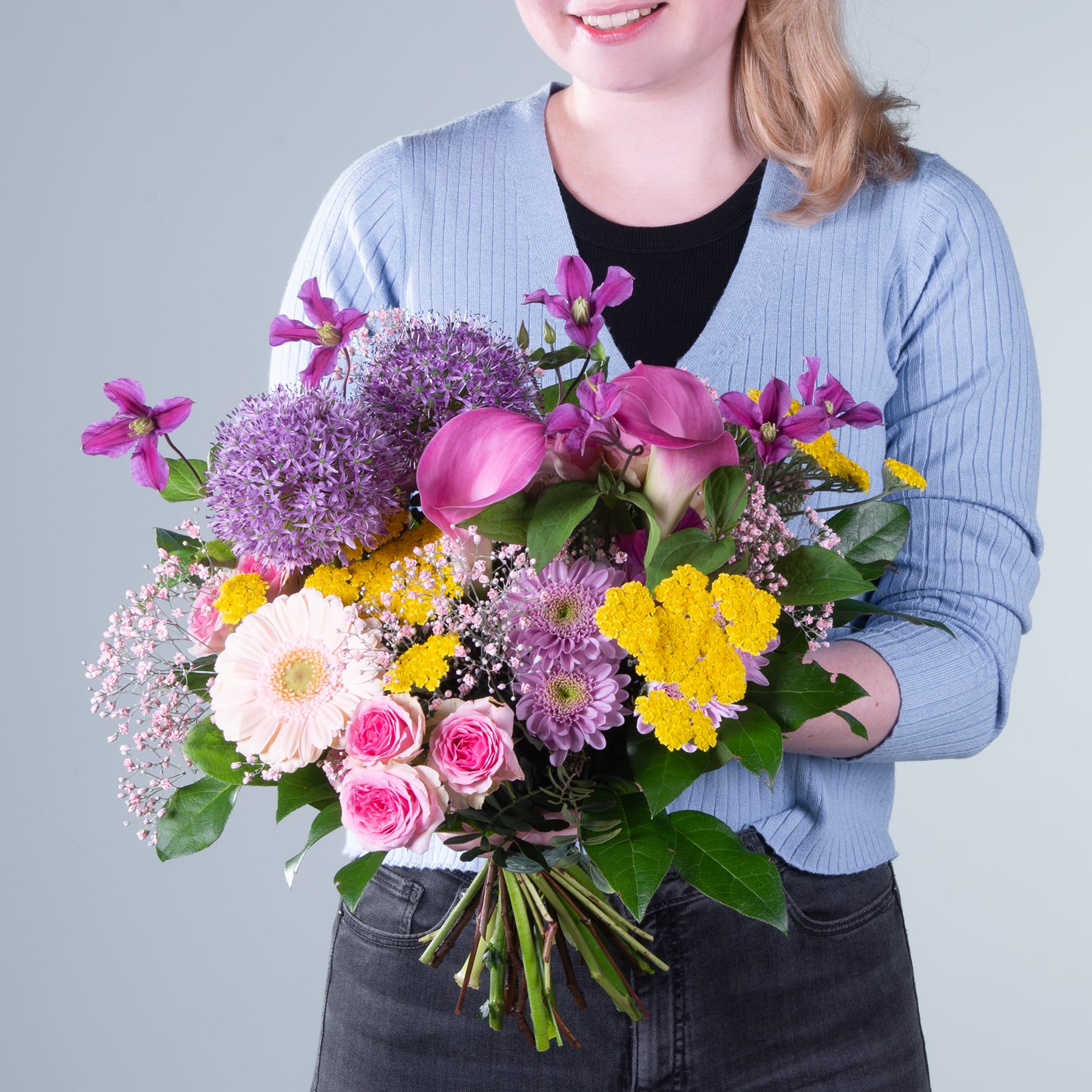 Eine Frau in einem grauen Pullover hält vor einem schlichten grauen Hintergrund einen leuchtend bunten Blumenstrauß, darunter rosa Rosen, lila Allium und gelbe Gänseblümchen.
