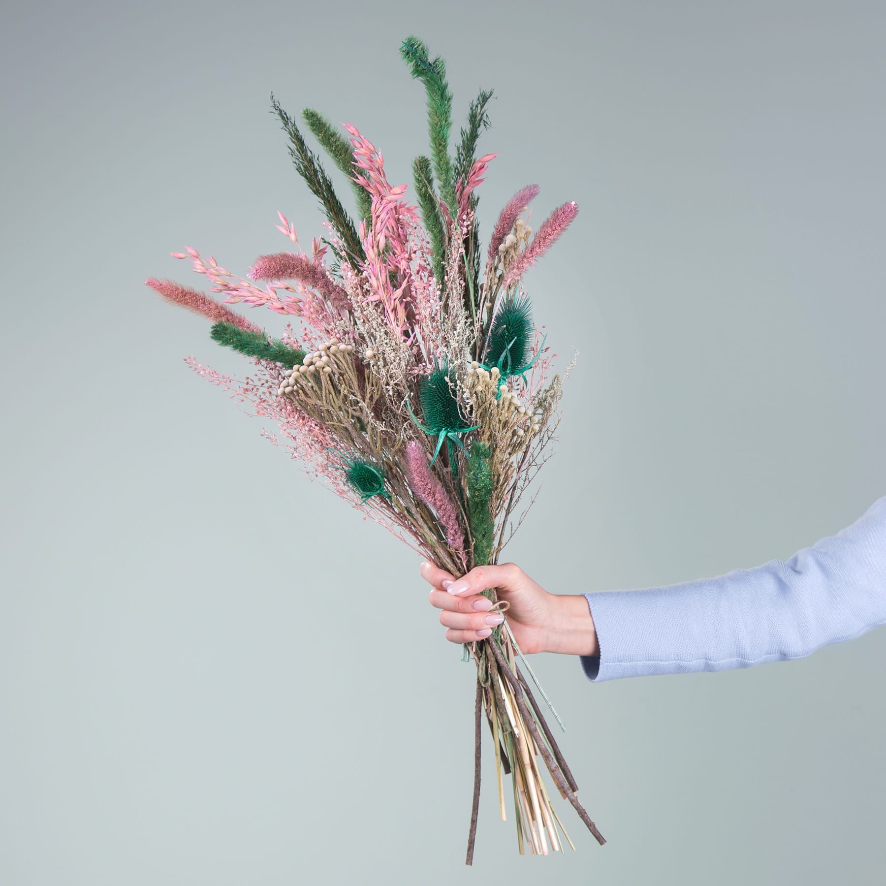 Eine Hand hält einen farbenfrohen Strauß getrockneter Blumen und Gräser in verschiedenen Rosa-, Grün- und Beigetönen vor einem schlichten hellgrauen Hintergrund.