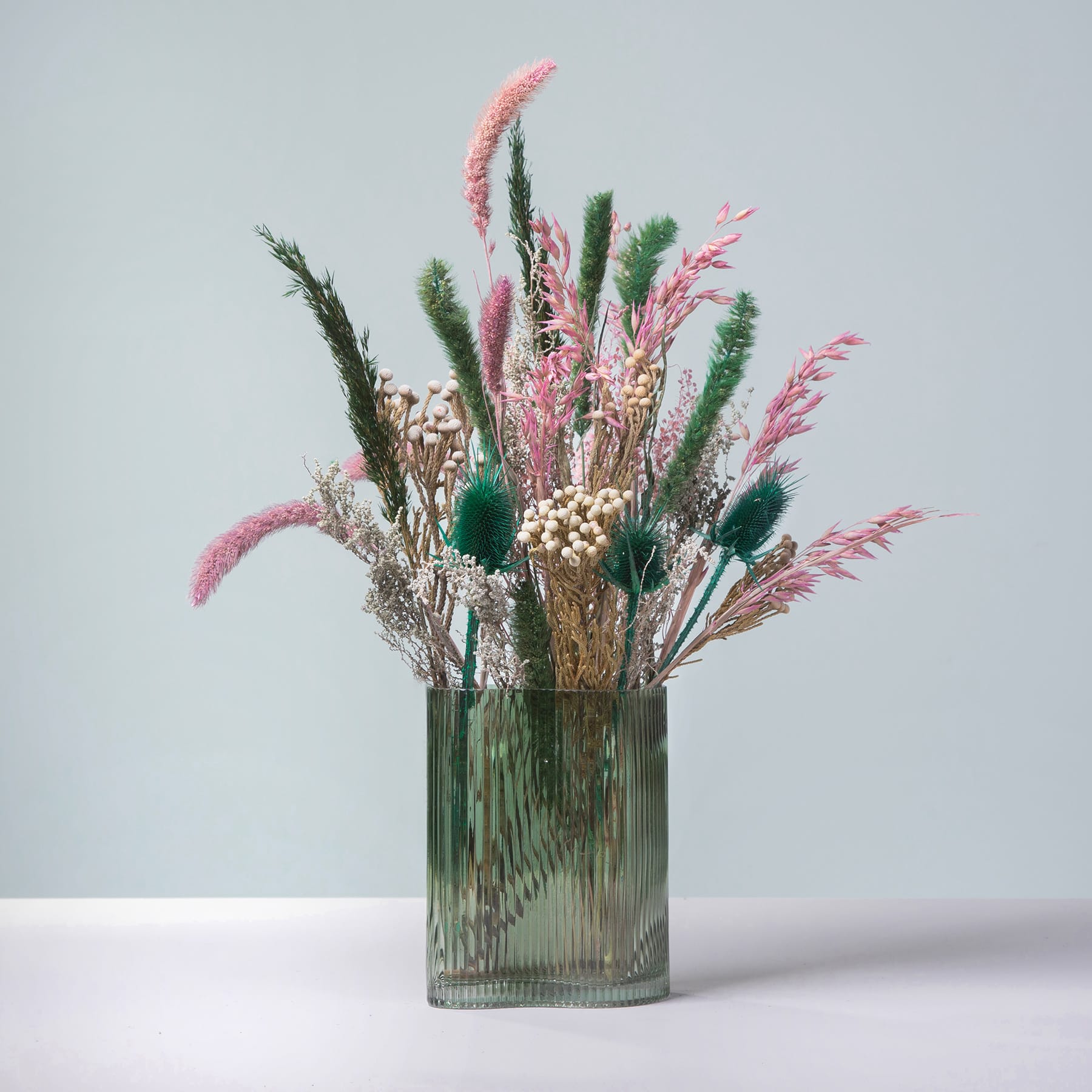 Eine Vase mit einem farbenfrohen Arrangement aus getrockneten Blumen und Grünpflanzen vor einem schlichten, hellen Hintergrund.