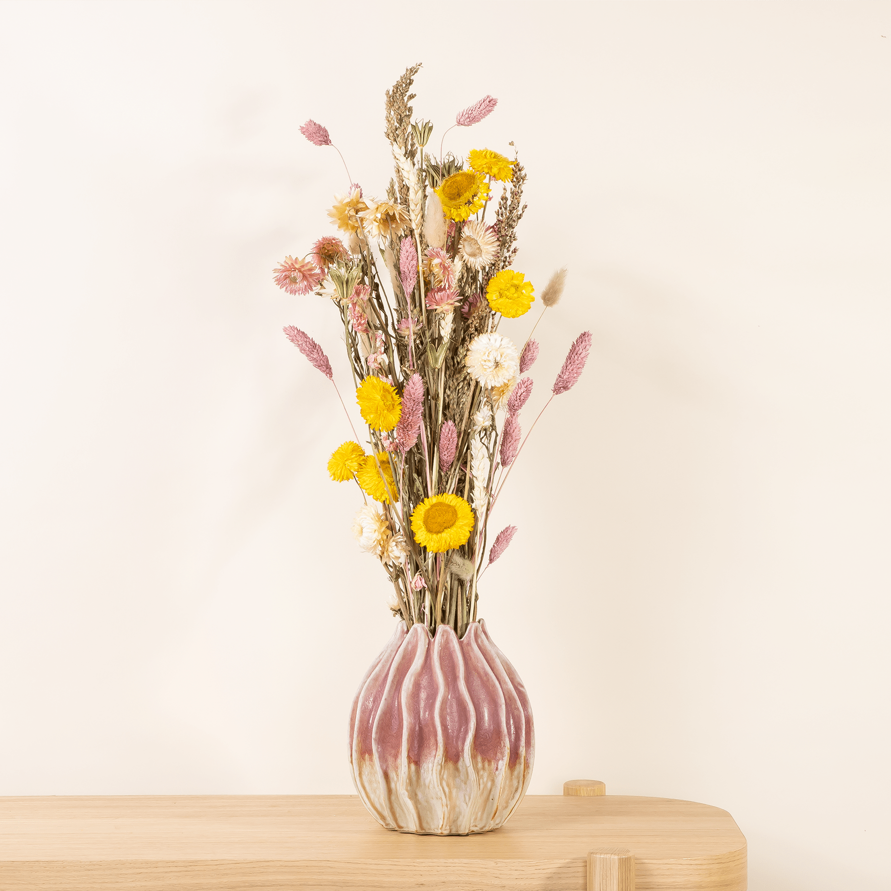 Ein farbenfroher Strauß getrockneter Blumen, darunter gelbe Sonnenblumen und rosa Blütenblätter, wird in einer dekorativen Keramikvase auf einer Holzoberfläche vor einem schlichten Hintergrund präsentiert.