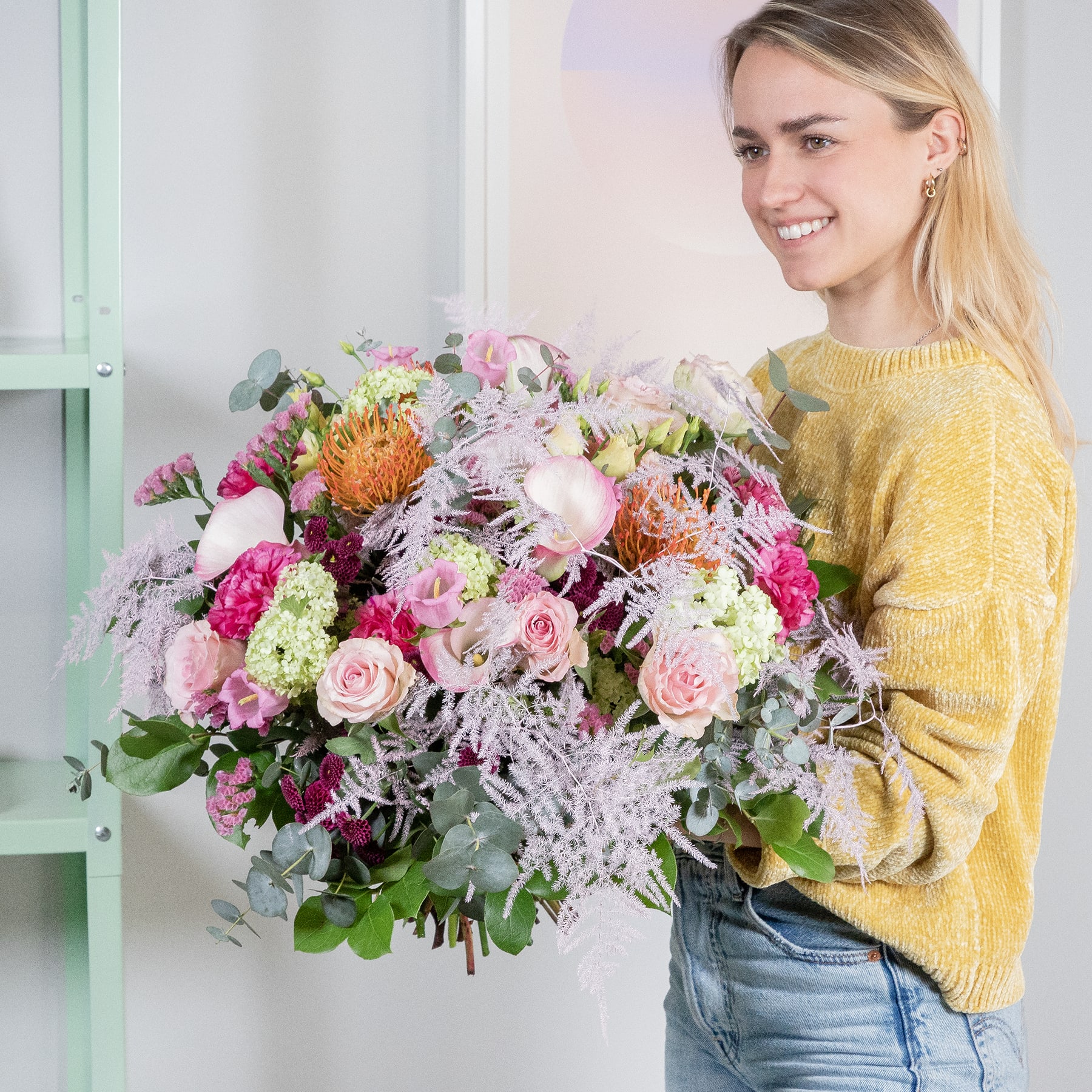 Eine lächelnde junge Frau in einem gelben Pullover hält einen großen, leuchtenden Strauß bunter Blumen vor einem hellen Hintergrund.