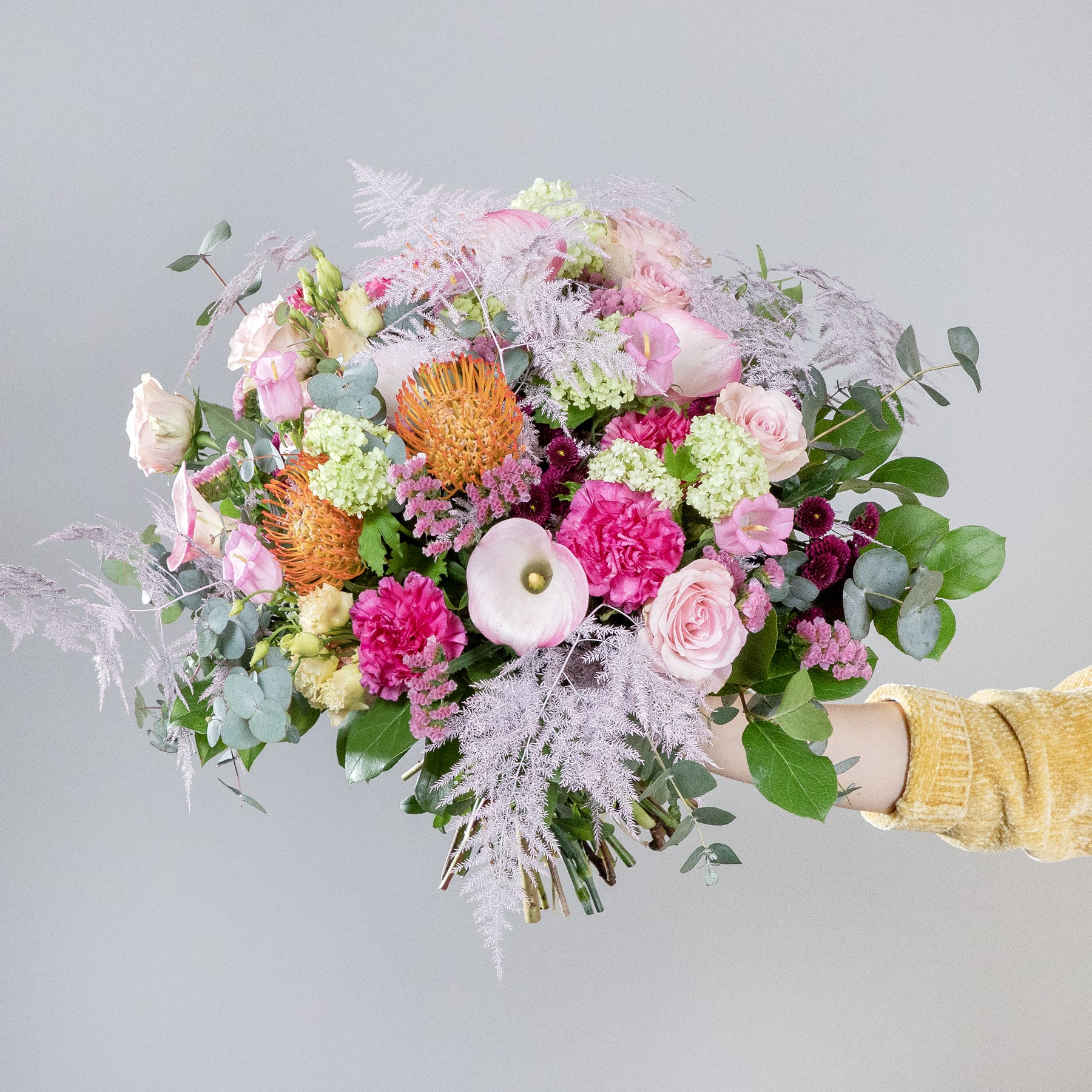 Ein lebendiger und farbenfroher Strauß verschiedener Blumen, darunter Pfingstrosen, Dahlien und Eukalyptus, den eine Person vor einem schlichten grauen Hintergrund in der Hand hält.