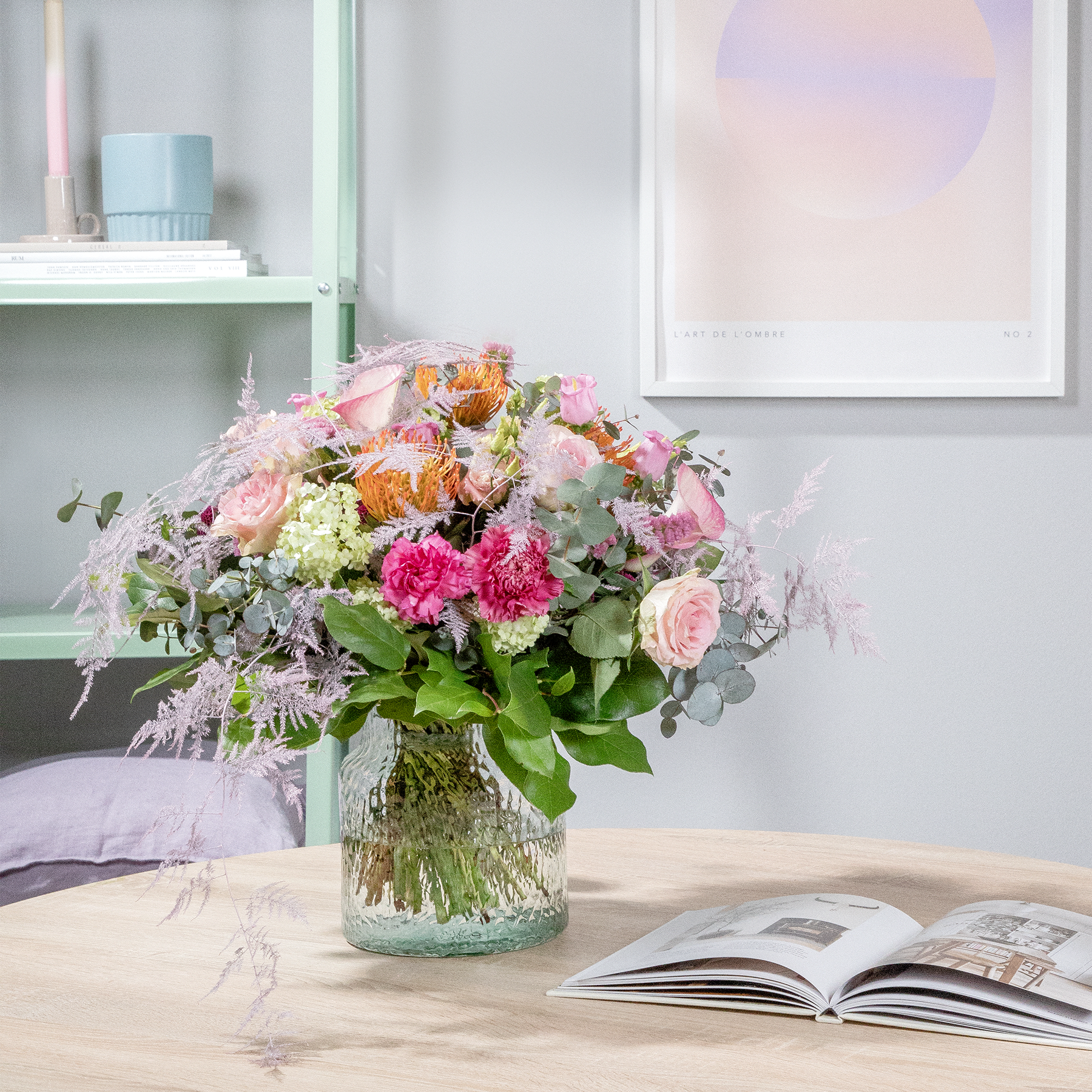 Ein farbenfroher Strauß rosa, gelber und weißer Blumen steht auf einem Holztisch vor einem großen Fenster, im Hintergrund sind ein Buch und andere Dekorationsgegenstände zu sehen.