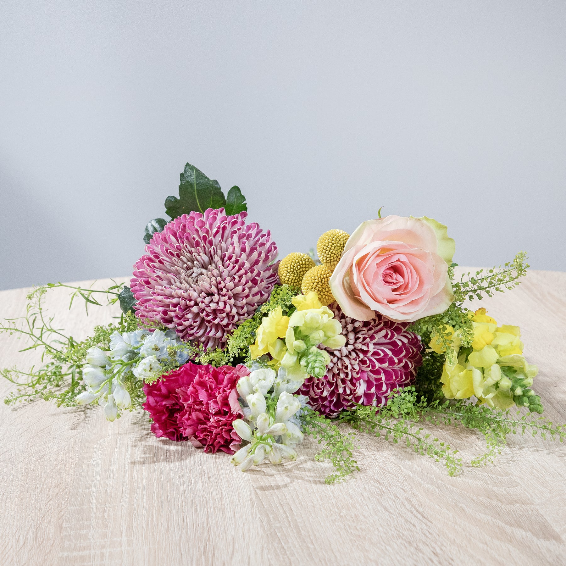 Ein farbenfroher Strauß rosa, gelber und weißer Blumen, darunter Rosen, Chrysanthemen und Grünpflanzen, liegt auf einer Holzoberfläche vor einem schlichten Hintergrund.