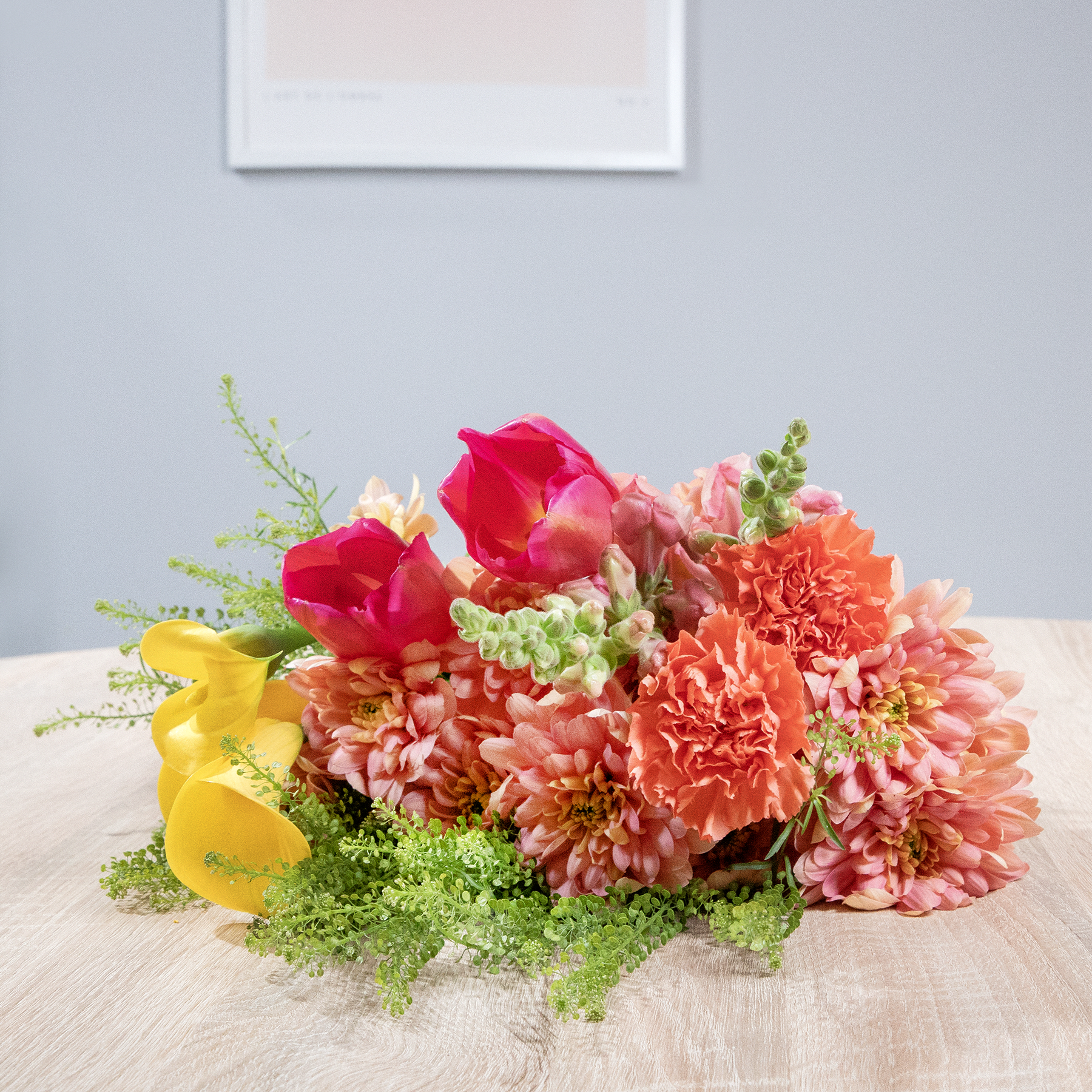 Ein farbenfroher Strauß roter Rosen, rosa Nelken und grünem Laub steht auf einem Holztisch vor einer weiß gerahmten Wand.
