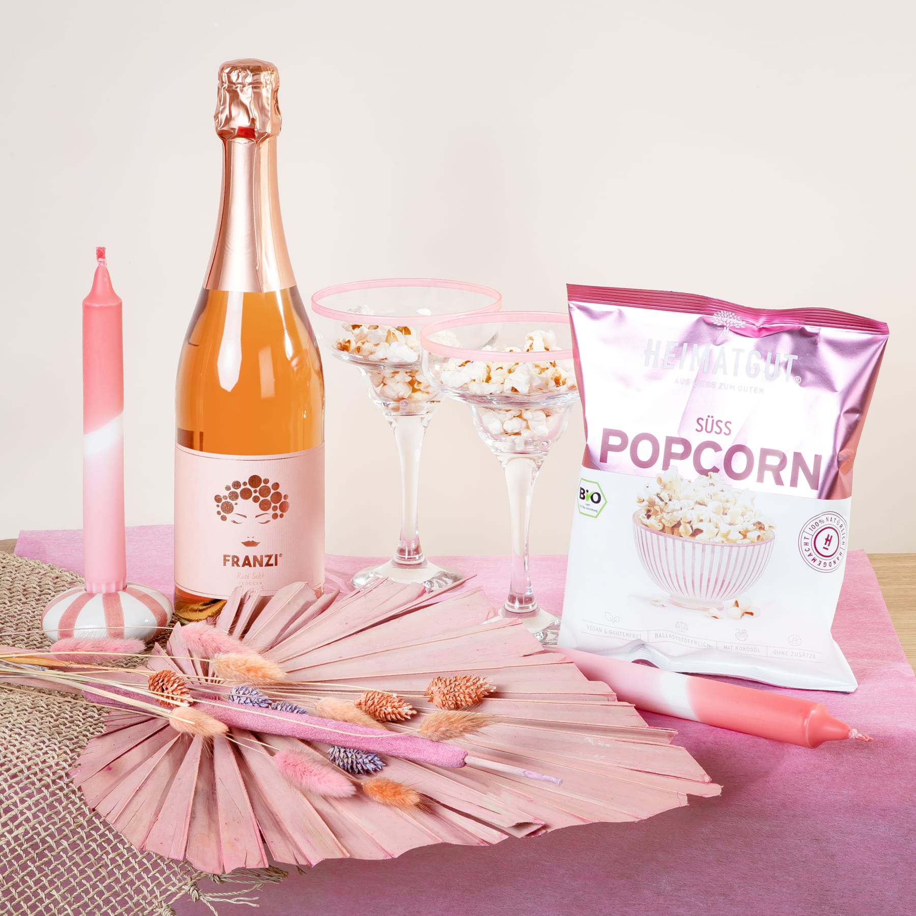 Ein festliches Arrangement mit einer Flasche Sekt, Popcorn und verschiedenen rosa und weißen Dekorationselementen auf einer strukturierten Oberfläche.