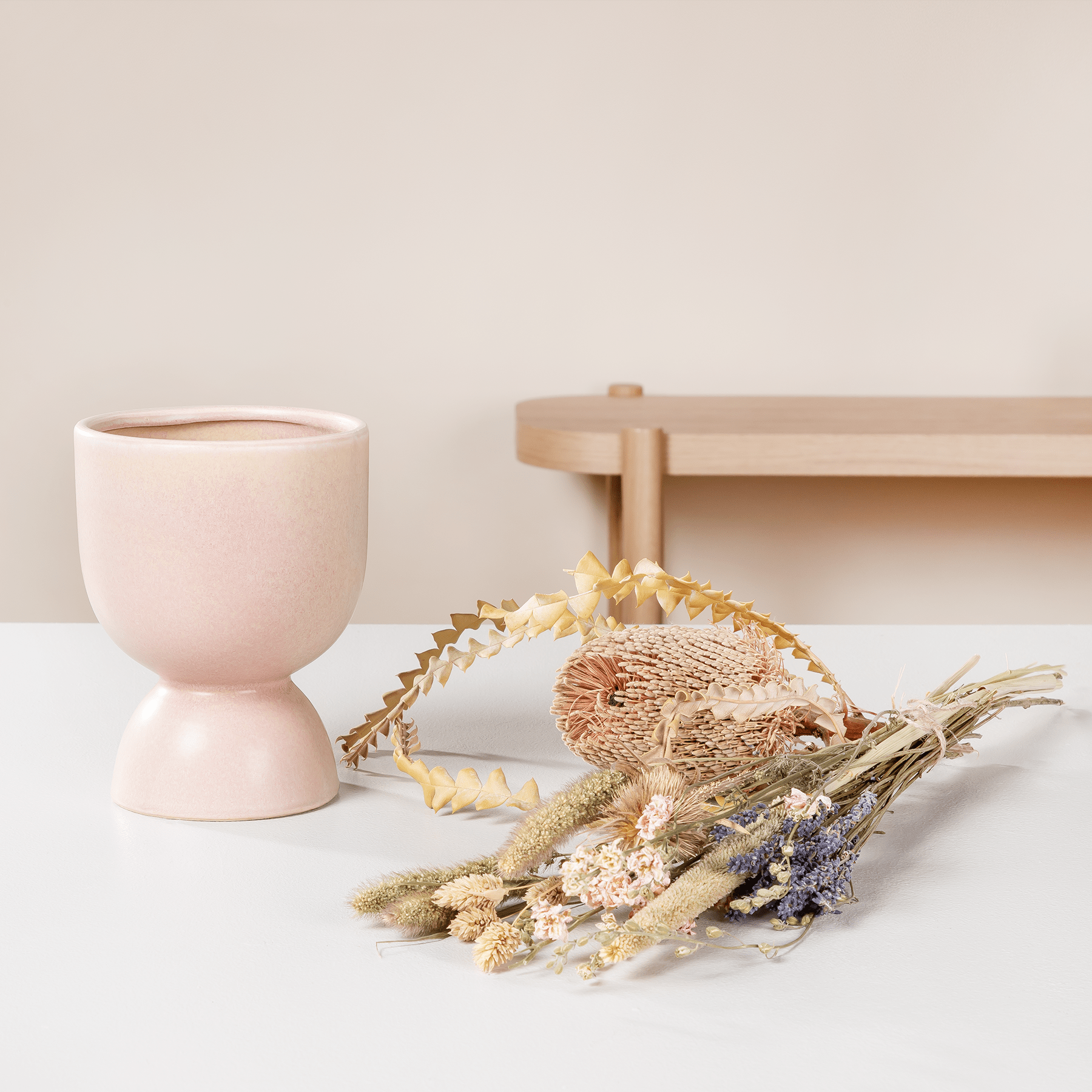 Ein Eierbecher aus Keramik in zartem Rosa, ein Holzkreuz und ein Strauß getrockneter Blumen und Pflanzen stehen auf einer hellen Oberfläche vor einem schlichten Hintergrund.