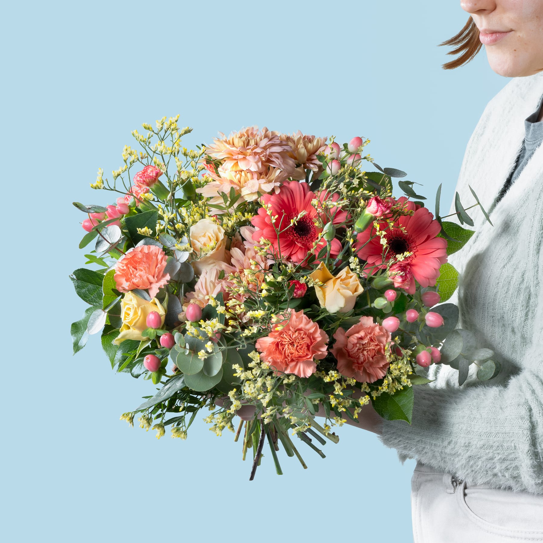 Ein leuchtender Strauß bunter Blumen, darunter Dahlien, Nelken und andere Blüten, wird von einer Person gehalten, die einen weißen Pullover vor einem hellblauen Hintergrund trägt.