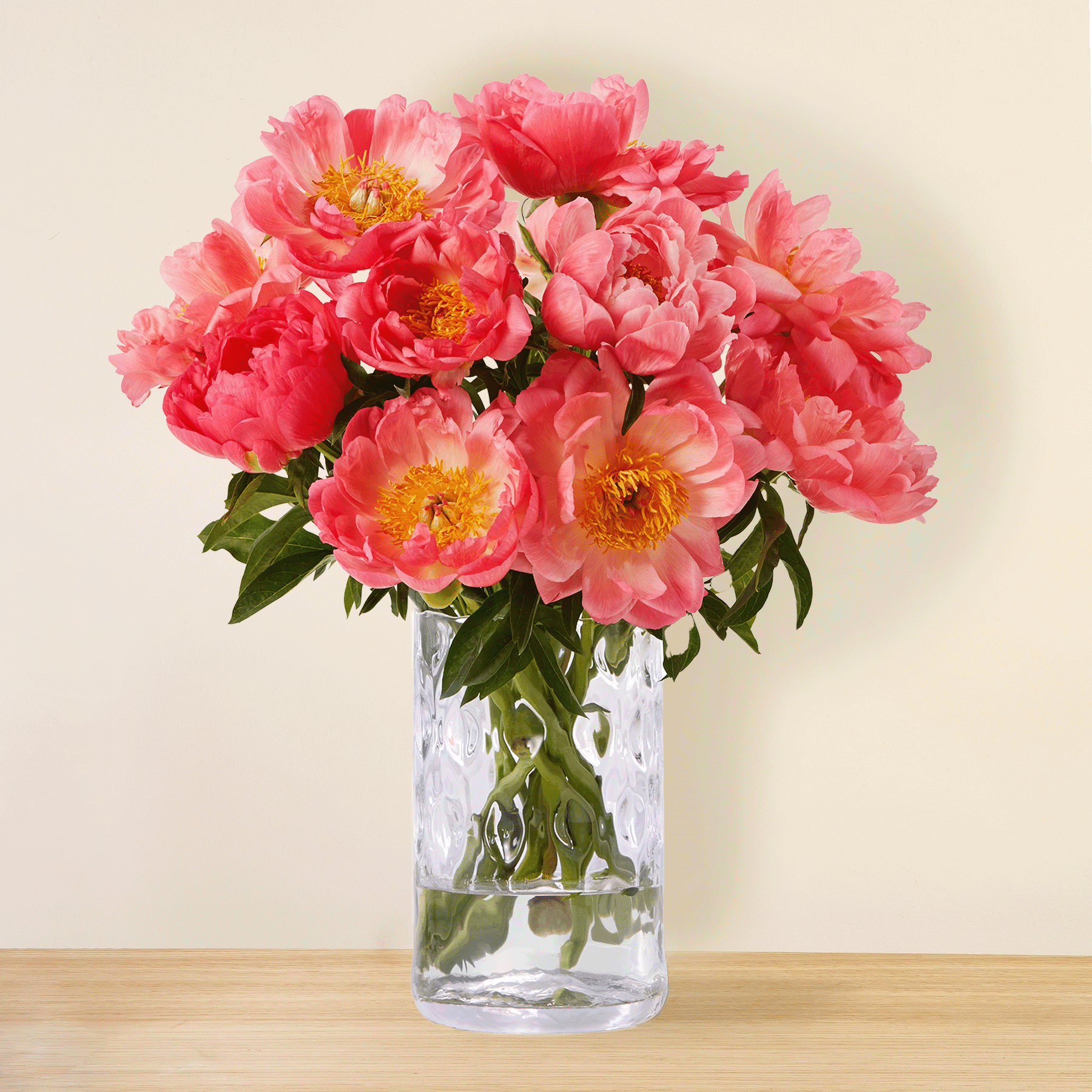 Ein Strauß leuchtend rosa Pfingstrosen in einer durchsichtigen Glasvase steht auf einer Holzoberfläche vor einem schlichten beigen Hintergrund.