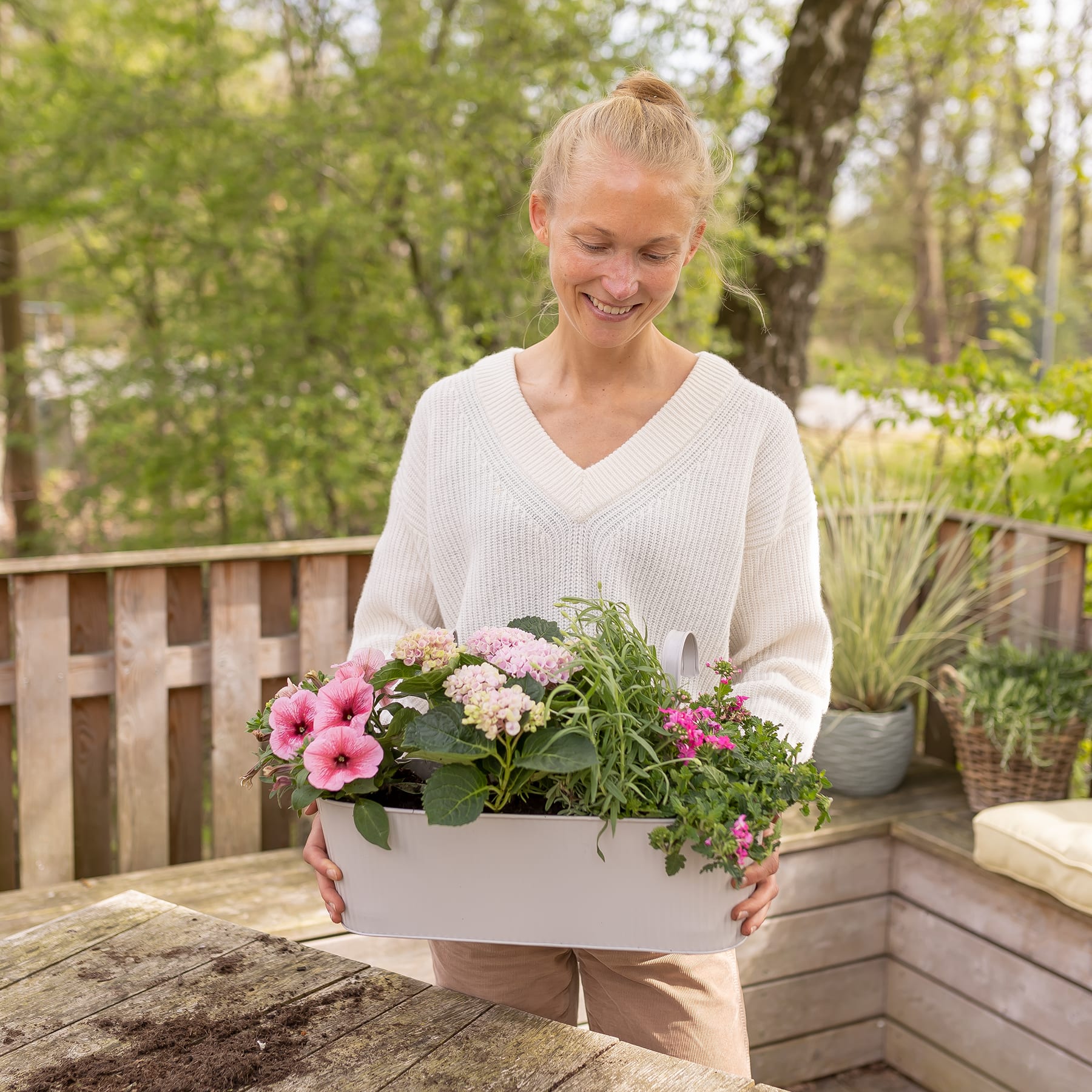 Eine lächelnde Frau in einem weißen Pullover hält einen Blumentopf voller bunter Blumen und Grünpflanzen und steht auf einem Holzdeck, umgeben von üppigem Laub und Bäumen.