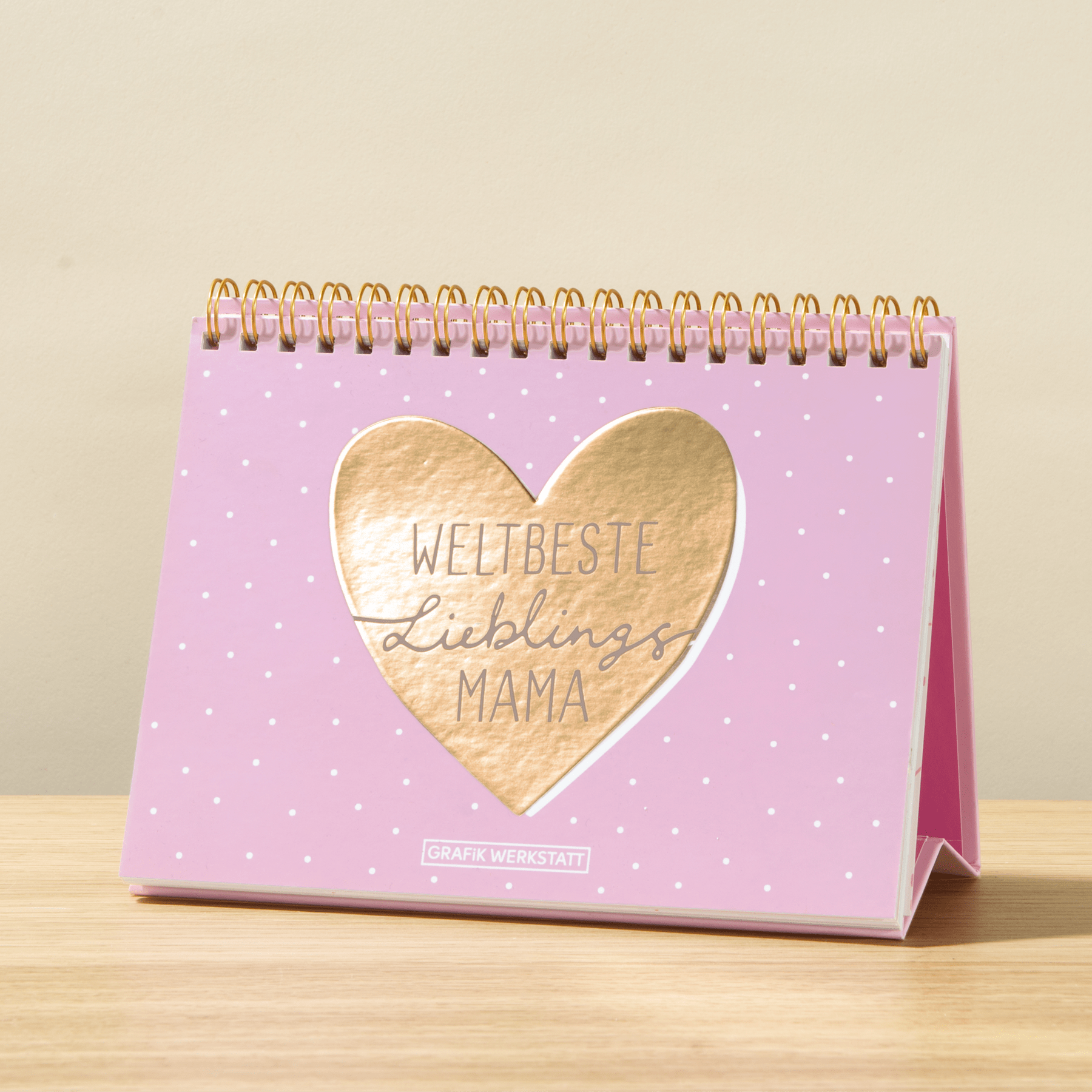Ein rosa Tischkalender mit Spiralbindung und einem herzförmigen Ausschnitt auf dem Cover, auf dem der Text „Weltbeste Lieblingsmama“ zu sehen ist.