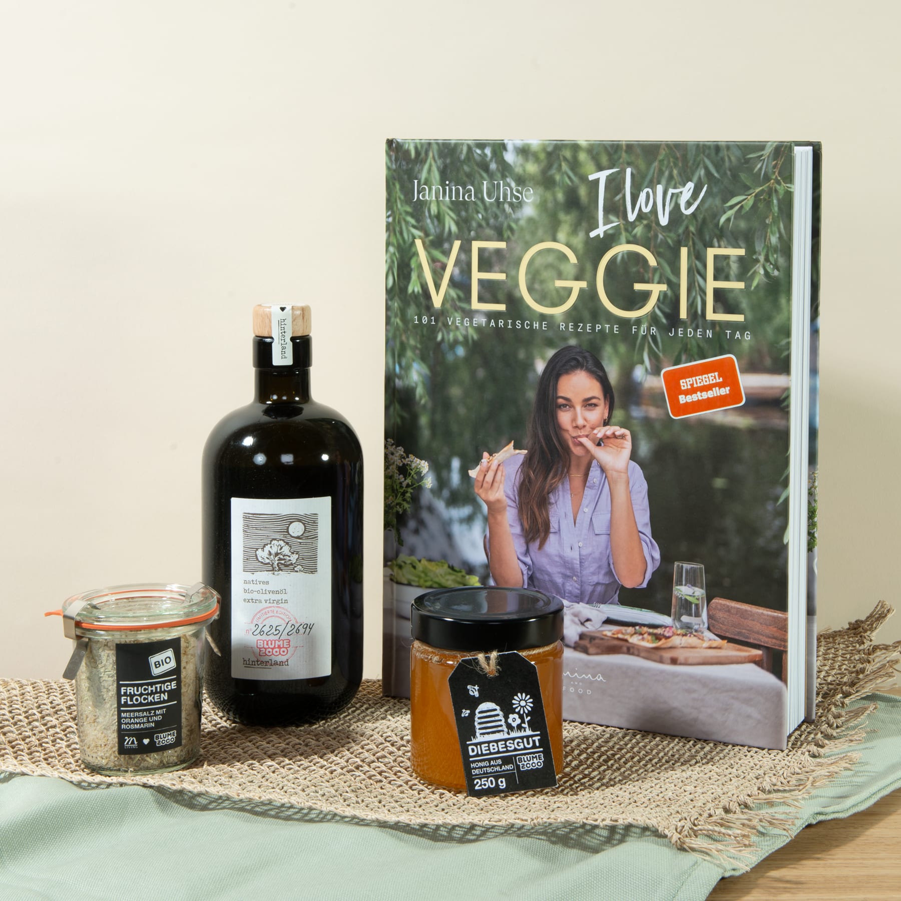 Das Bild zeigt ein Kochbuch mit dem Titel „I Love Veggie“ von Katrina Ells, zusammen mit einer Flasche Wein und einem Glas mit etwas, das wie Honig oder ein ähnliches Gewürz aussieht, alles auf einer gewebten Matte oder Tischdecke platziert.