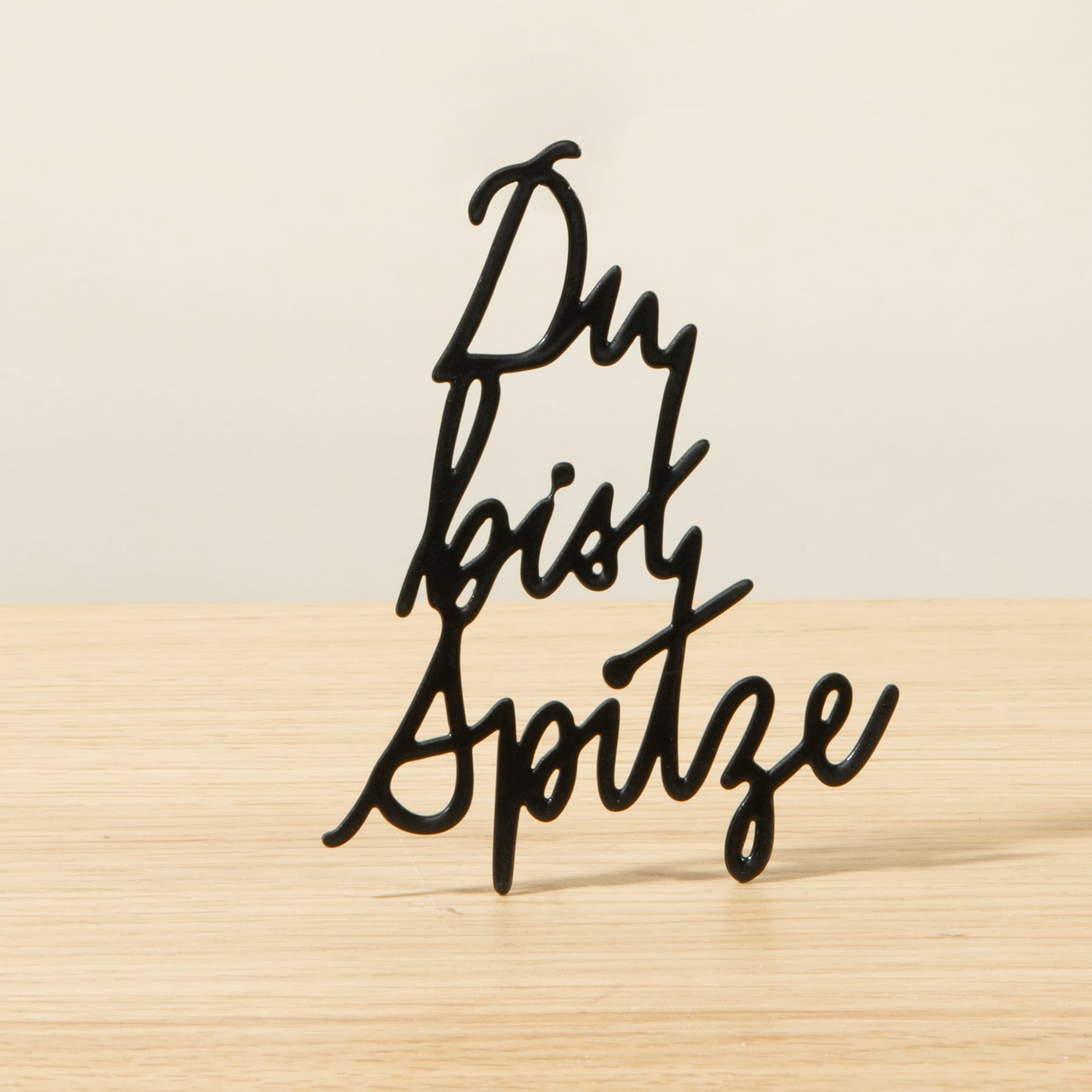 Das Bild zeigt den Text „Du bist Spitze“ in einer stilisierten, handgeschriebenen Schriftart vor einem schlichten, hellen Holzhintergrund.