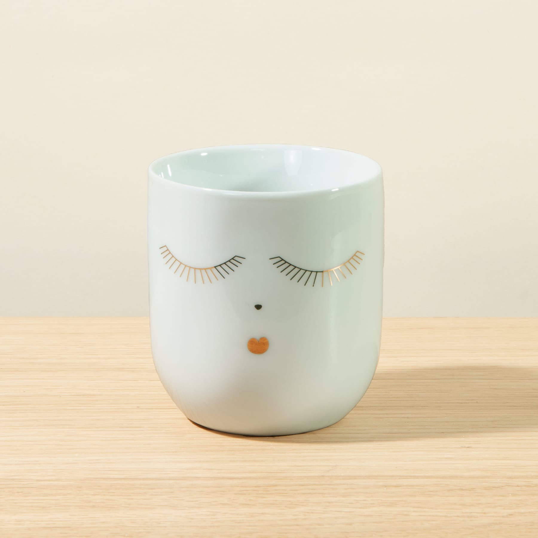 Eine weiße Keramiktasse oder ein Becher mit einem einfachen Gesichtsdesign, mit geschlossenen Augen und einem kleinen orangefarbenen Punkt als Nase, steht auf einer Holzoberfläche vor einem hellen Hintergrund.