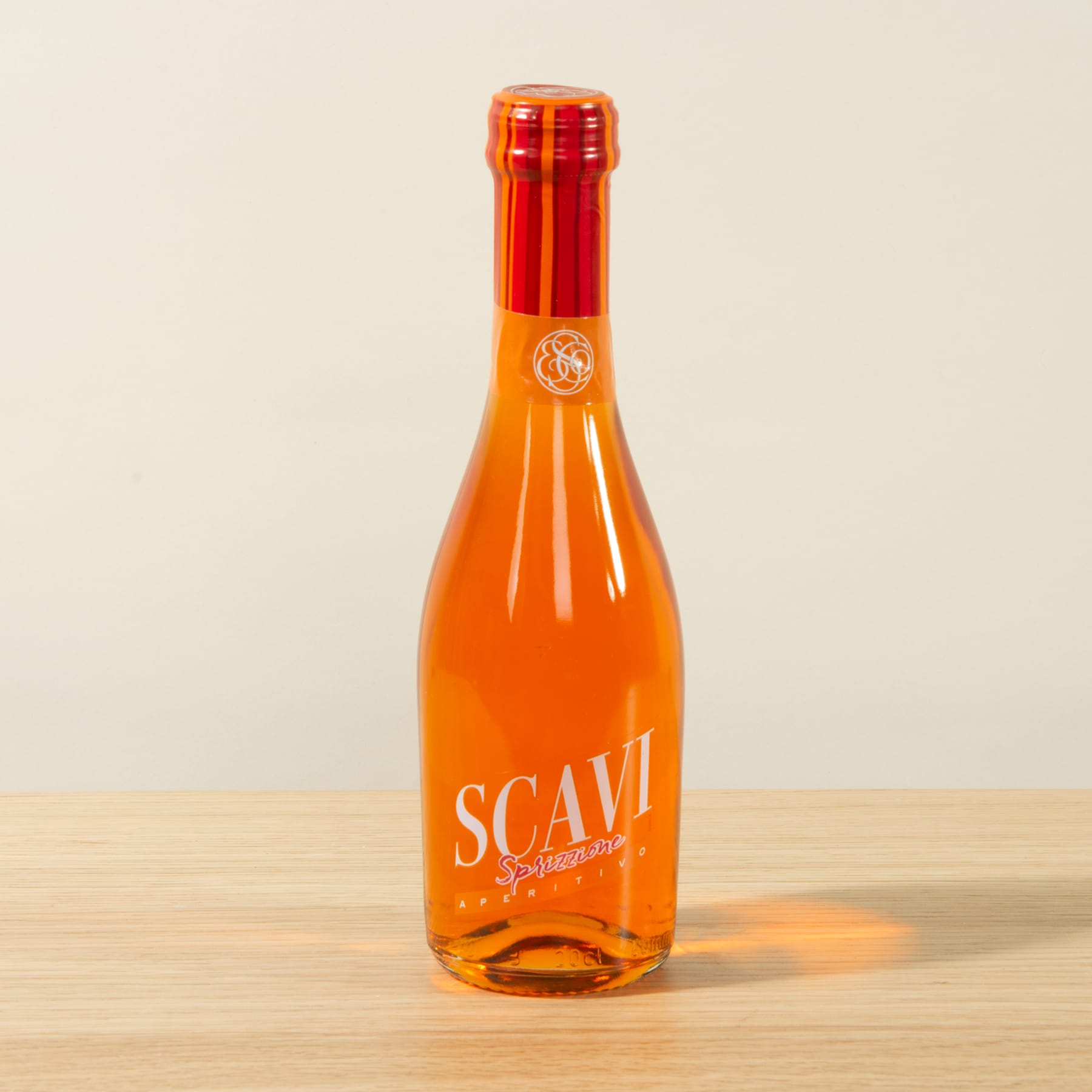 Eine leuchtend orangefarbene Glasflasche mit dem auffälligen Etikett „SCAVI“ steht auf einer Holzoberfläche vor einem schlichten Hintergrund.