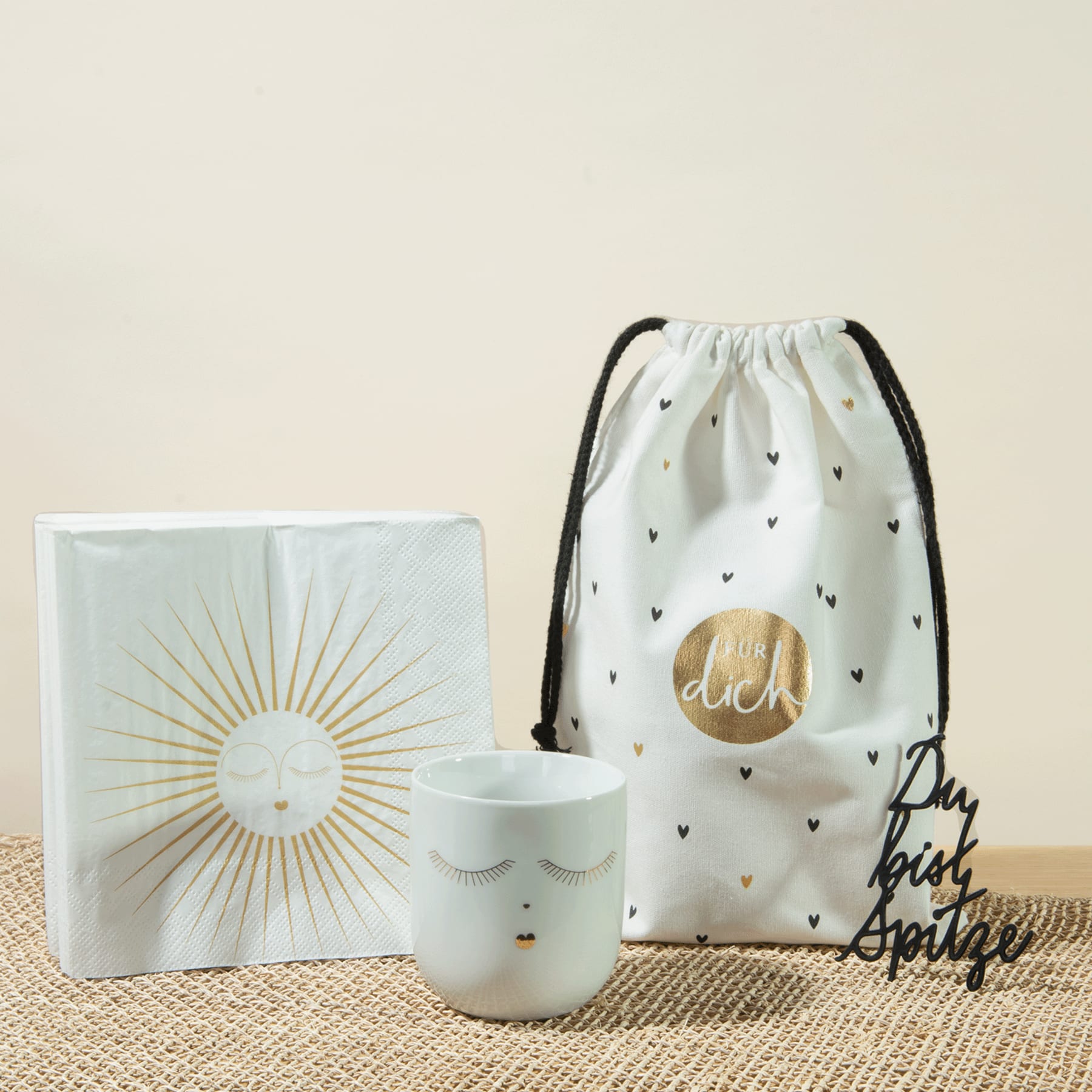 Auf einem beigen Untergrund sind ein weißer Kordelbeutel mit Sonnenmotiv, eine weiße Keramiktasse und eine weiße Box mit Sonnenmotiv angeordnet.