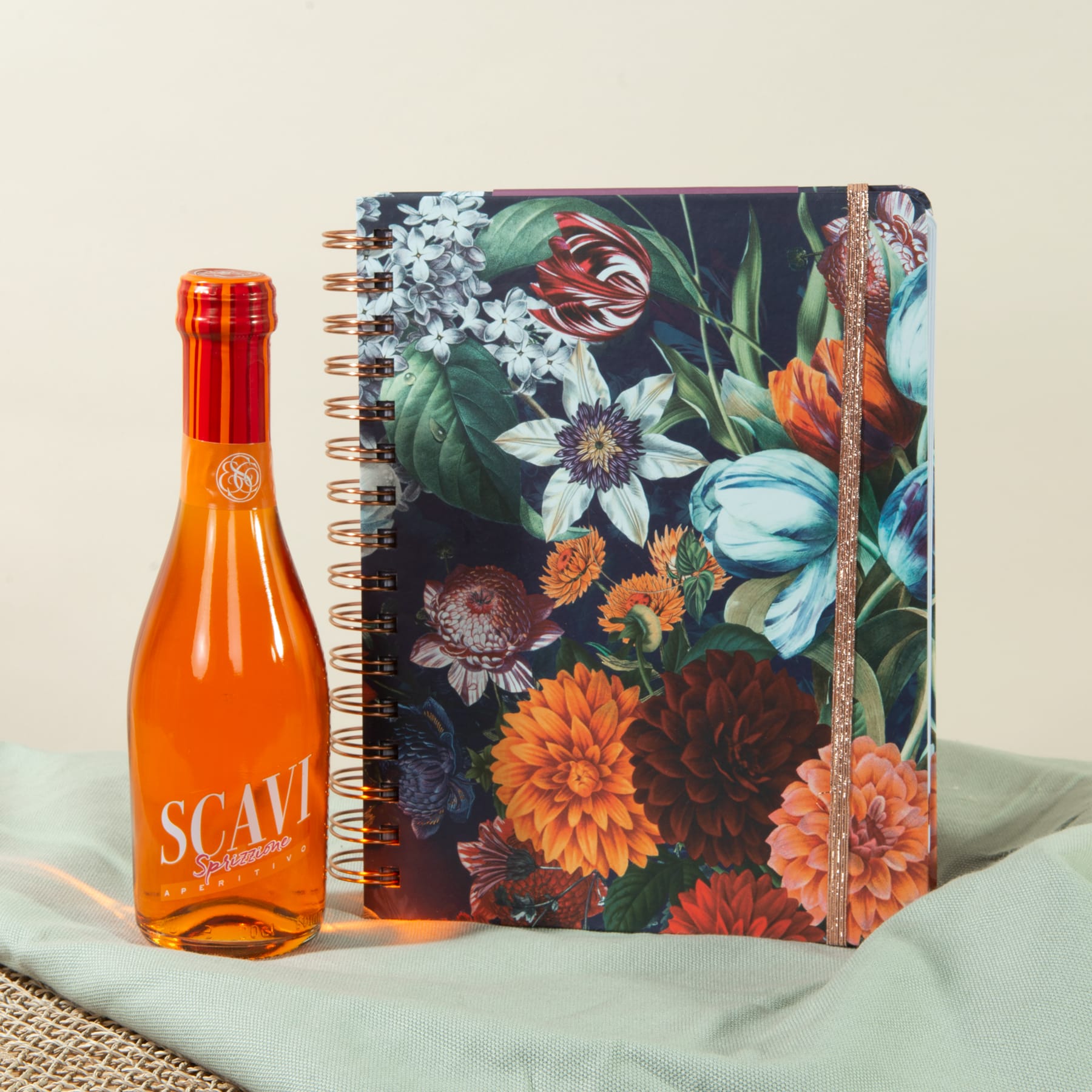 Auf einem hellen Stoffhintergrund sind ein farbenfrohes Notizbuch mit Blumenmuster und eine Flasche mit orangefarbener Flüssigkeit abgebildet.