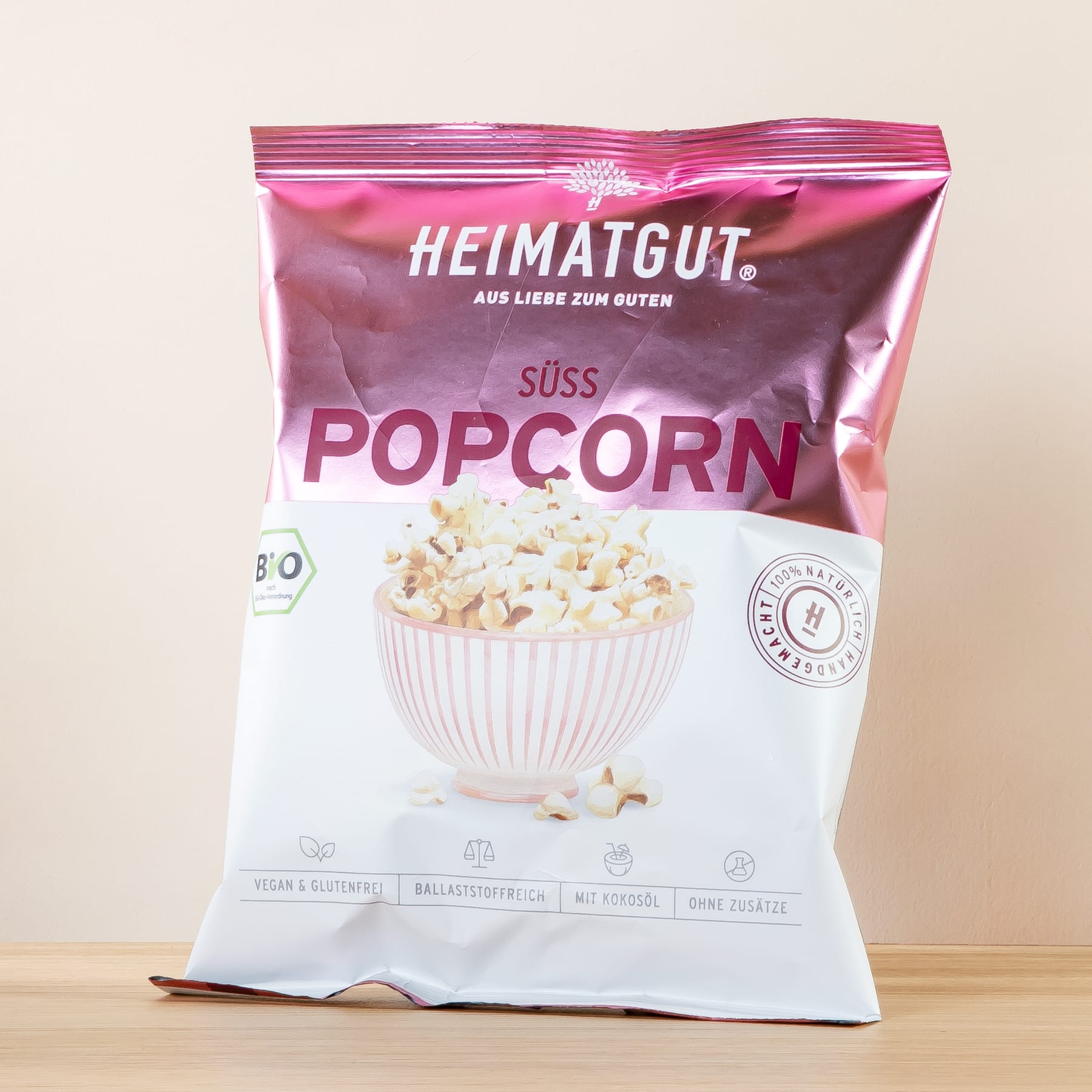 Auf einer Holzfläche liegt eine rosa-weiße Tüte süßes Popcorn der Marke Heimatgut.