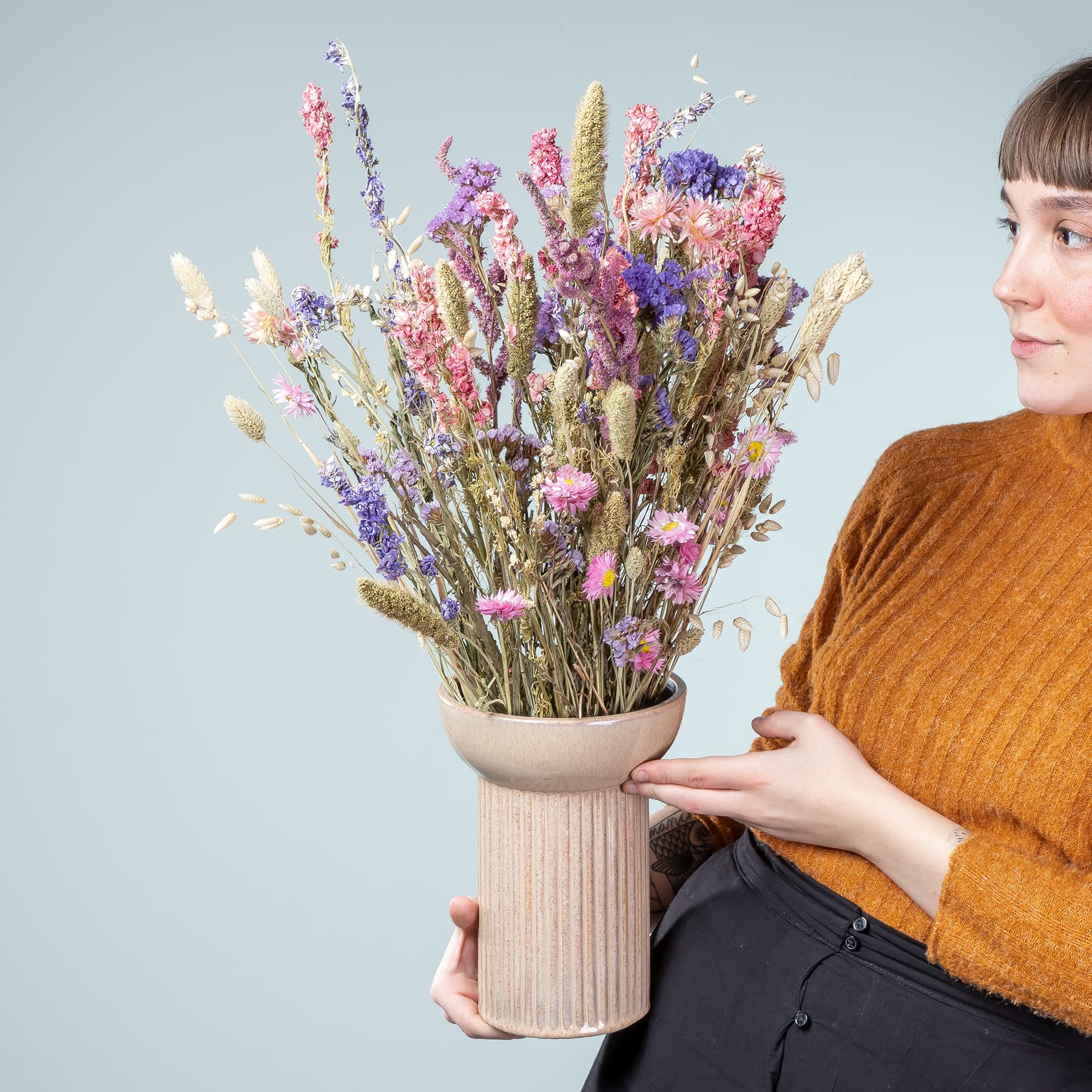 Eine Person in einem braunen Pullover hält vor einem schlichten blauen Hintergrund eine große Vase mit einem leuchtenden Strauß bunter Trockenblumen und Gräser.