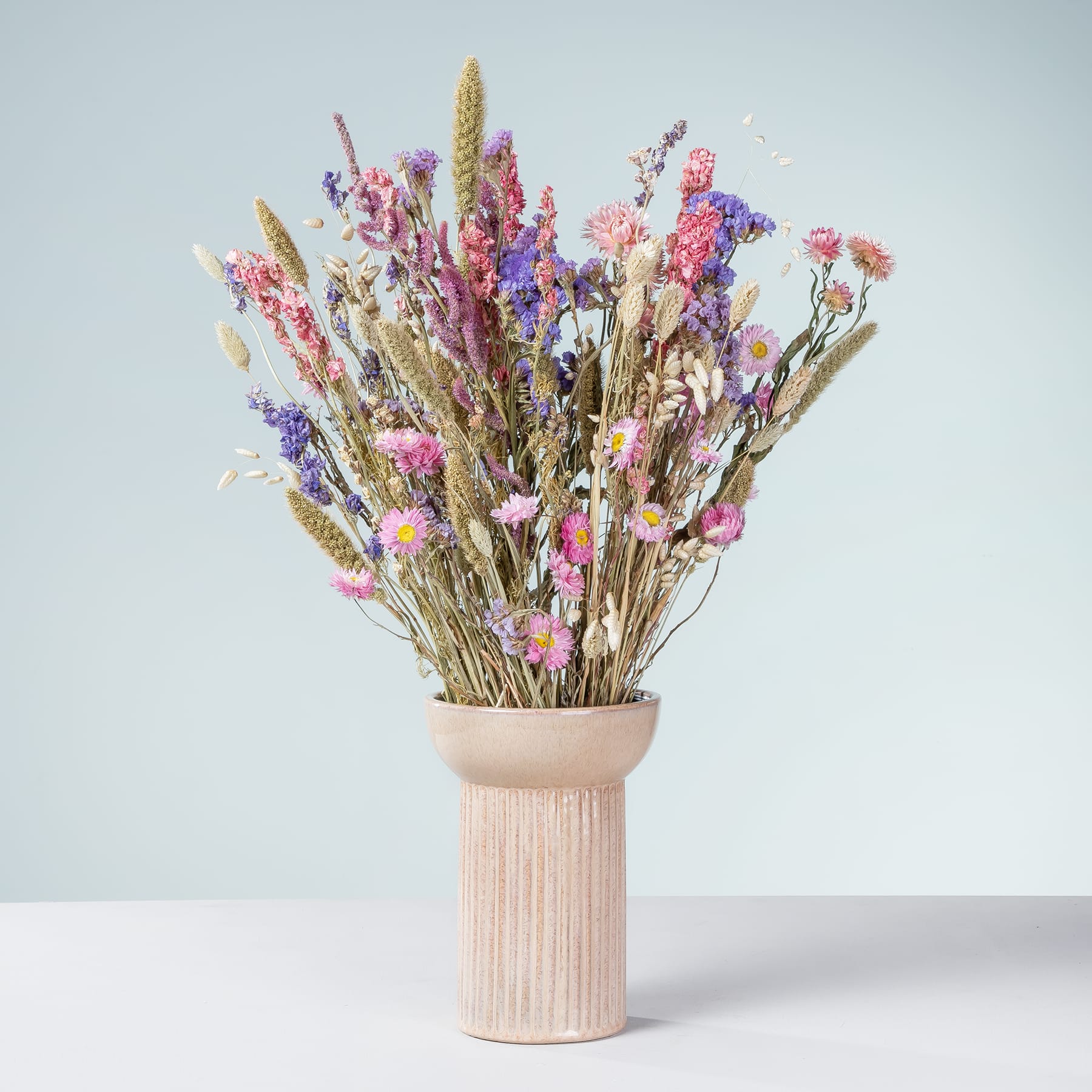 Ein Strauß leuchtender Trockenblumen, darunter in Rosa- und Lilatönen, steht hoch in einer einfachen Keramikvase vor einem schlichten hellblauen Hintergrund.