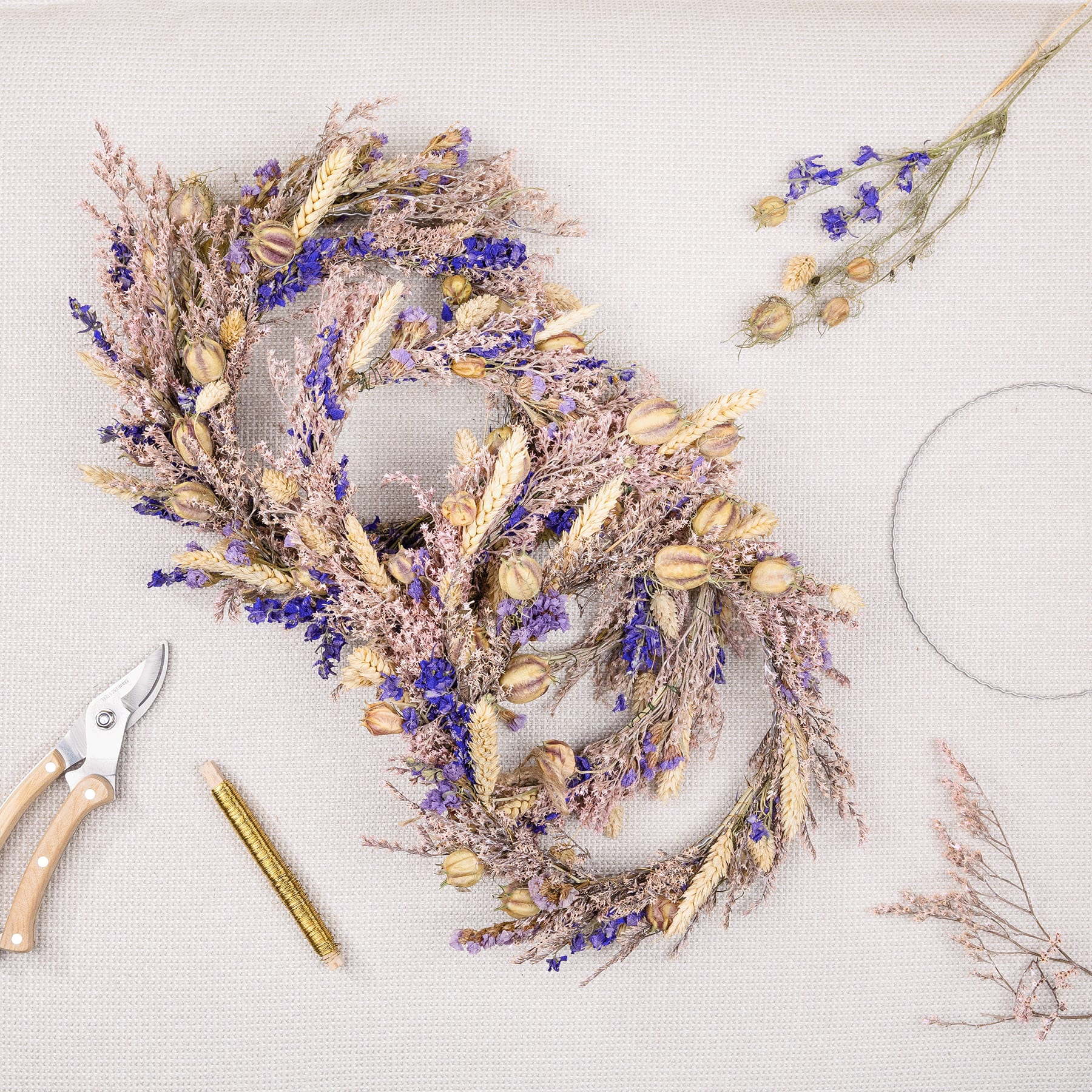 Auf einem hellen Hintergrund wird ein wunderschöner Kranz aus getrocknetem Lavendel, Weizen und anderen natürlichen Elementen präsentiert, begleitet von verschiedenen Gartengeräten und zusätzlichen floralen Elementen.