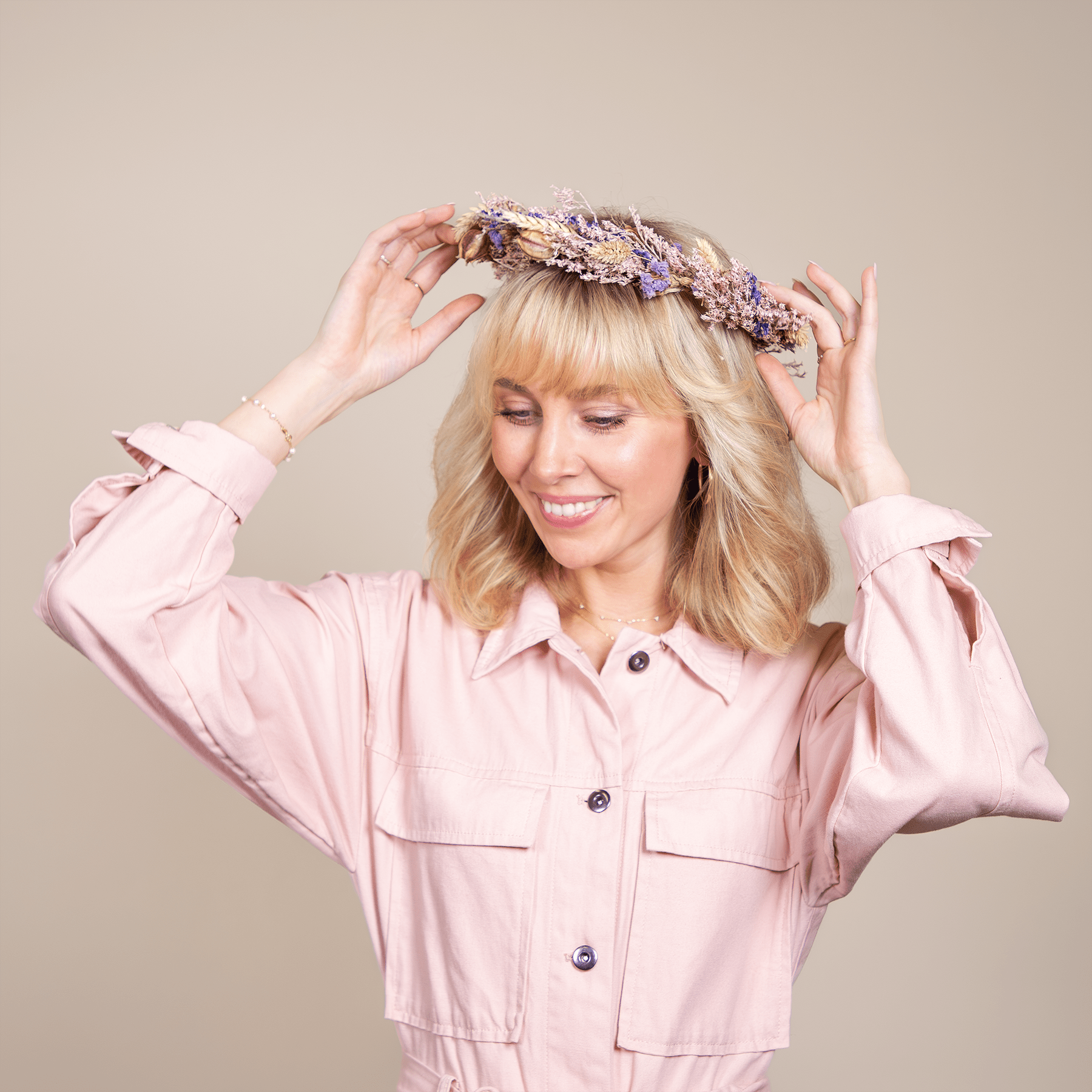Eine lächelnde Frau mit blondem Haar, einer rosa Jacke und einem Blumenkopfschmuck steht vor einem neutralen Hintergrund.